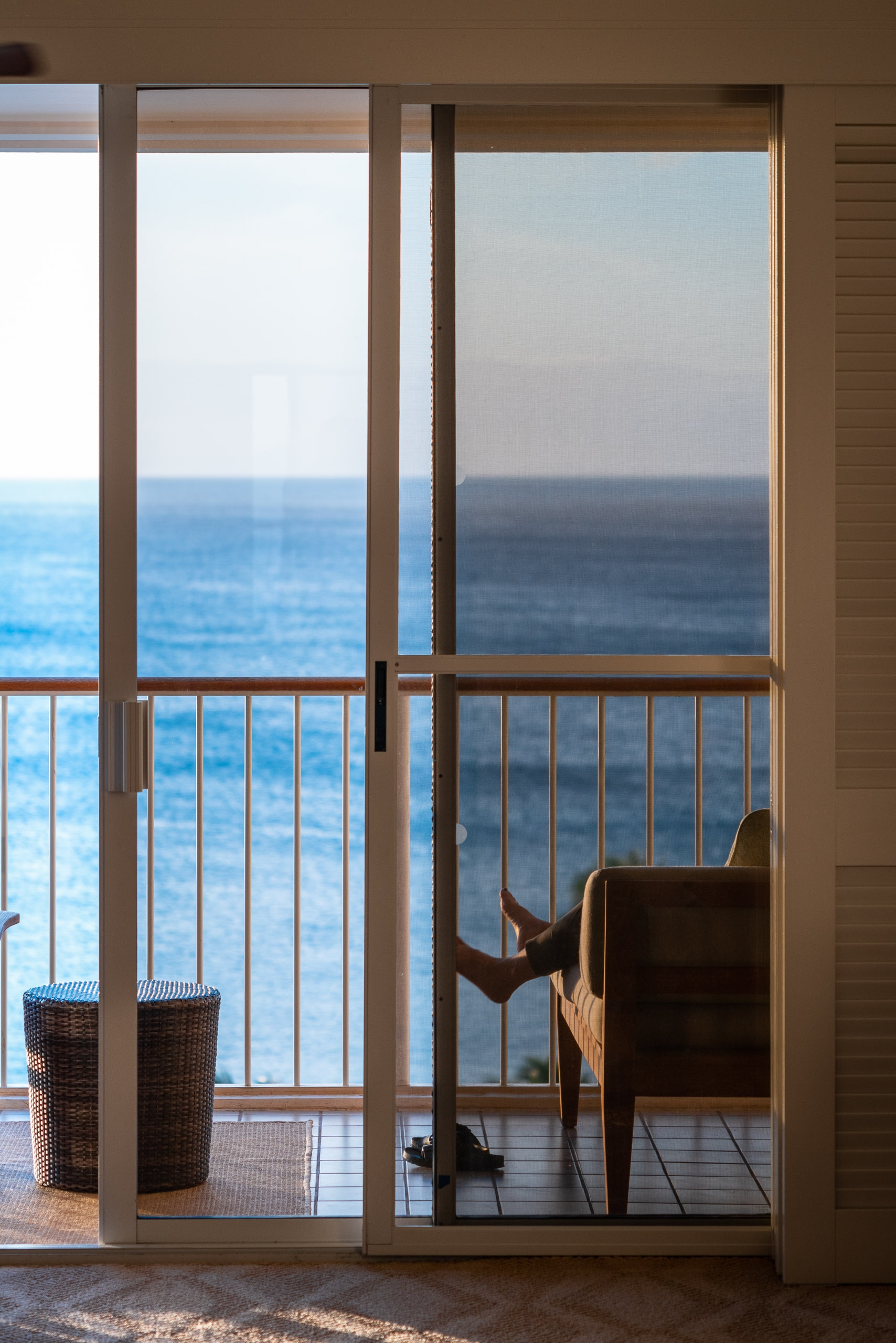 A beige interior room with glass sliding doors and metal railings opens onto a seaside balcony, revealing a dark wooden chair silhouetted against calm blue ocean waters beyond