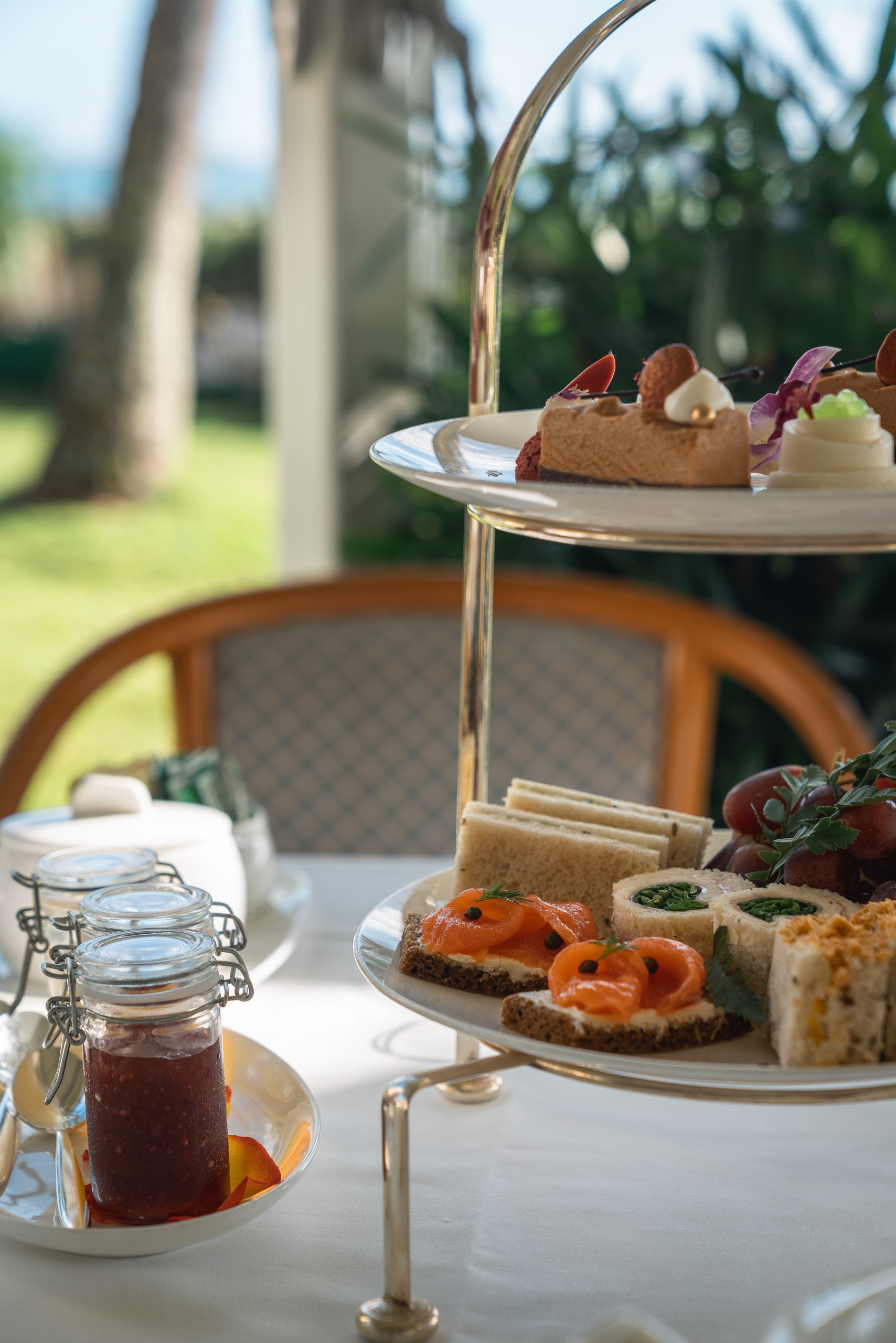 A tiered stand displays an elegant afternoon tea service with finger sandwiches, pastries, and fresh berries in a sunny garden setting with palm trees visible in the background