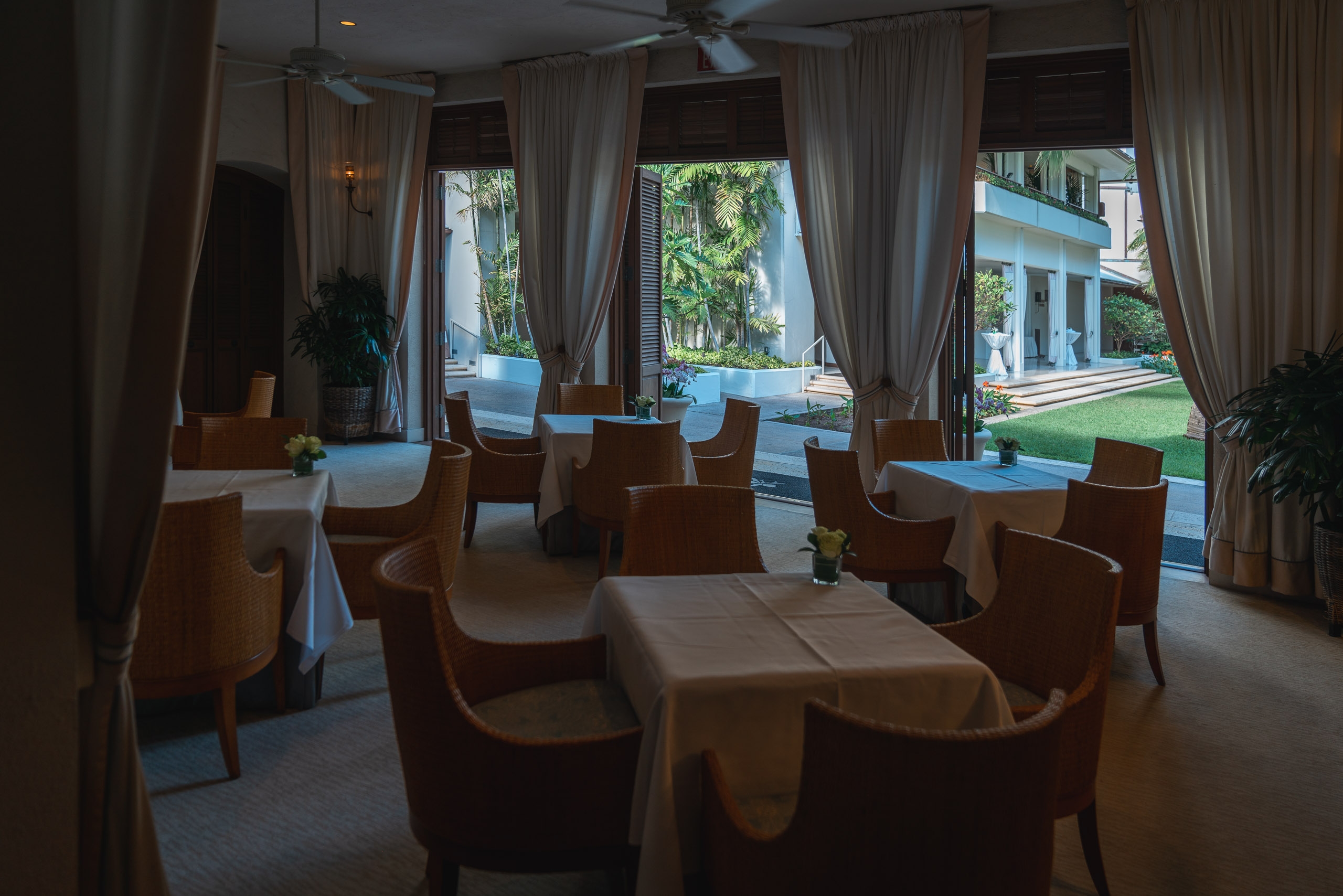 An elegant dining room with wooden chairs arranged around white-clothed tables overlooks a manicured lawn and tropical garden through expansive floor-to-ceiling windows framed by flowing beige curtains
