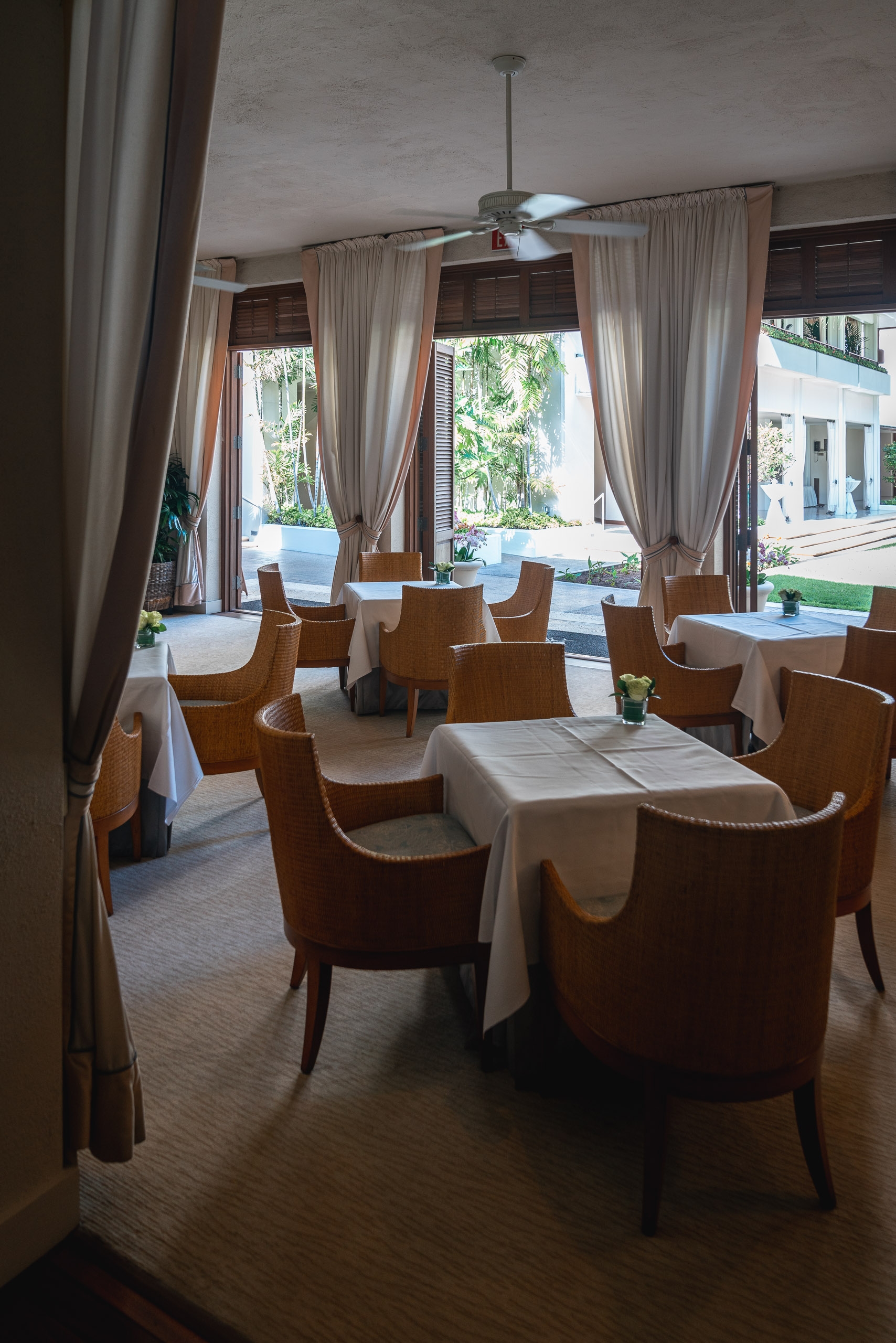 An upscale restaurant dining room features wood-framed chairs with white tablecloths, sheer curtains framing glass doors that open onto a sunny tropical courtyard with lush greenery and modern architecture visible beyond