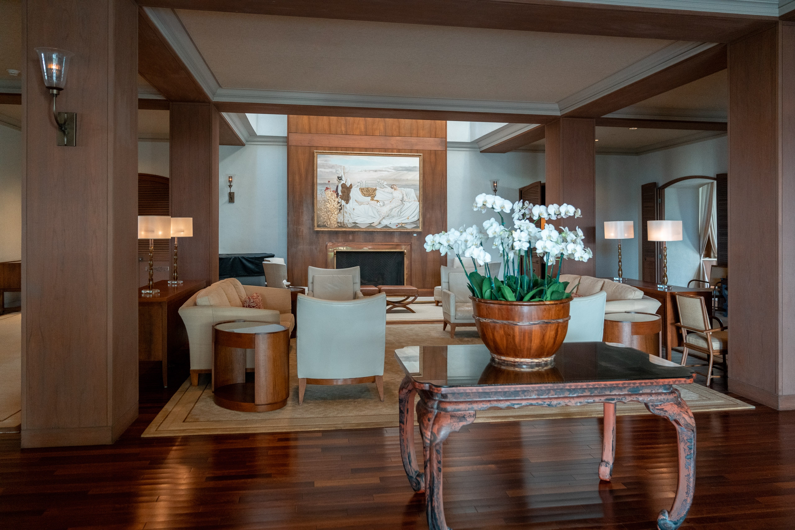 A sophisticated hotel lobby features rich wood paneling, cream-colored furniture, and a striking white orchid arrangement in a wooden vessel, illuminated by warm ambient lighting