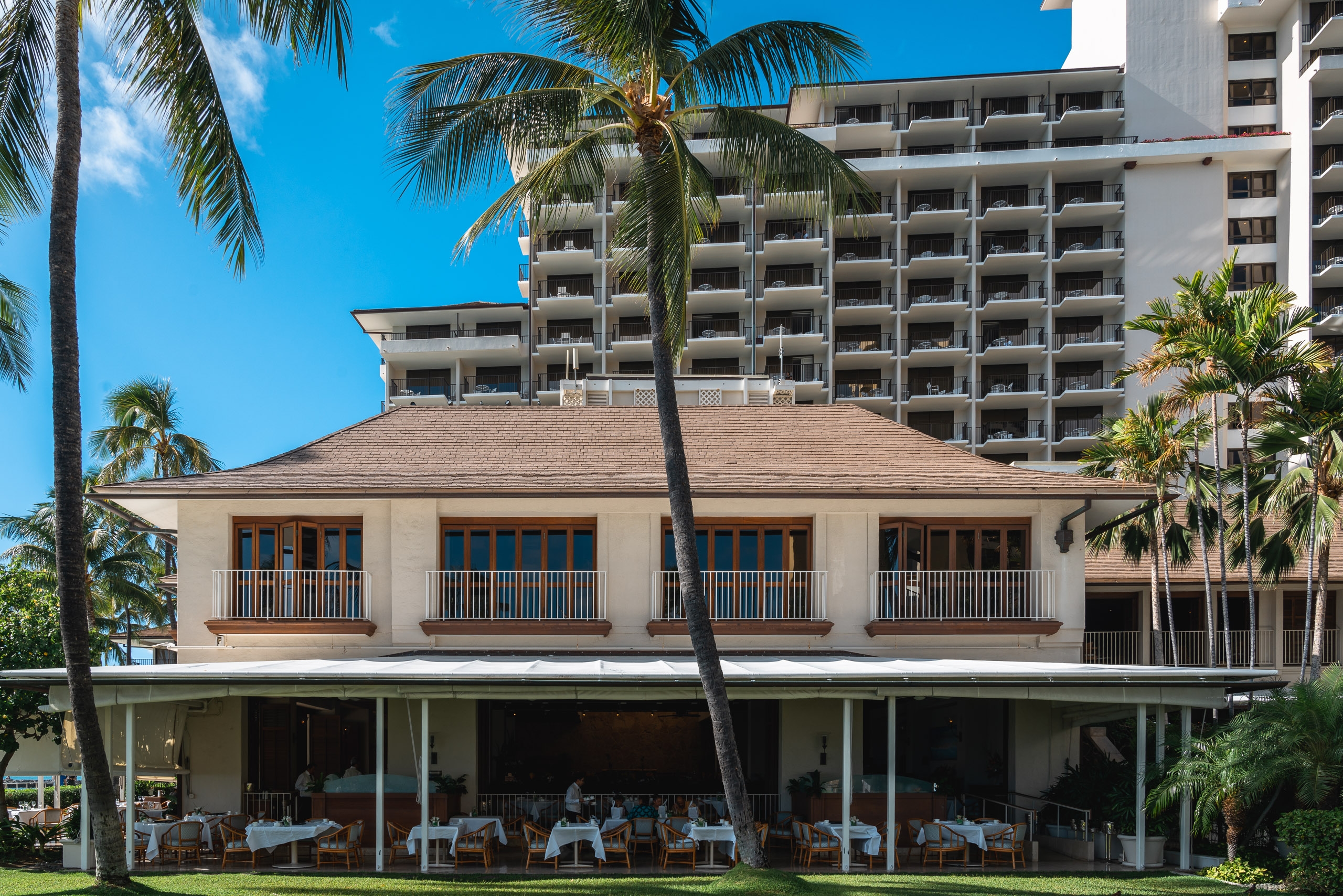 A historic beige hotel building with blue-trimmed windows and a wide ground-floor veranda overlooks manicured grounds, framed by palm trees against modern high-rise structures and clear blue sky