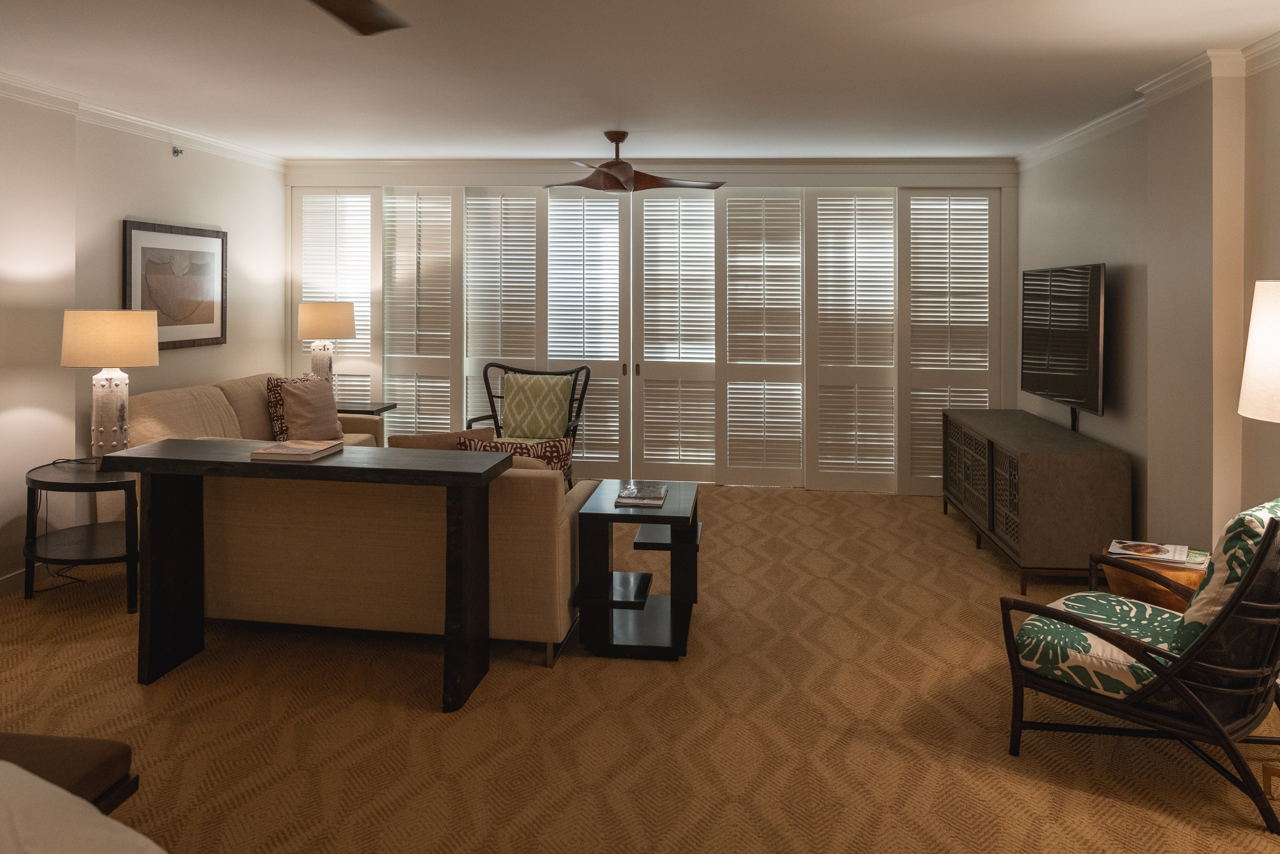 A spacious hotel suite with tan and brown furnishings, featuring a dark desk, lounge chairs, patterned carpeting, and full-length shuttered doors opening onto a lanai with soft ambient lighting throughout the room