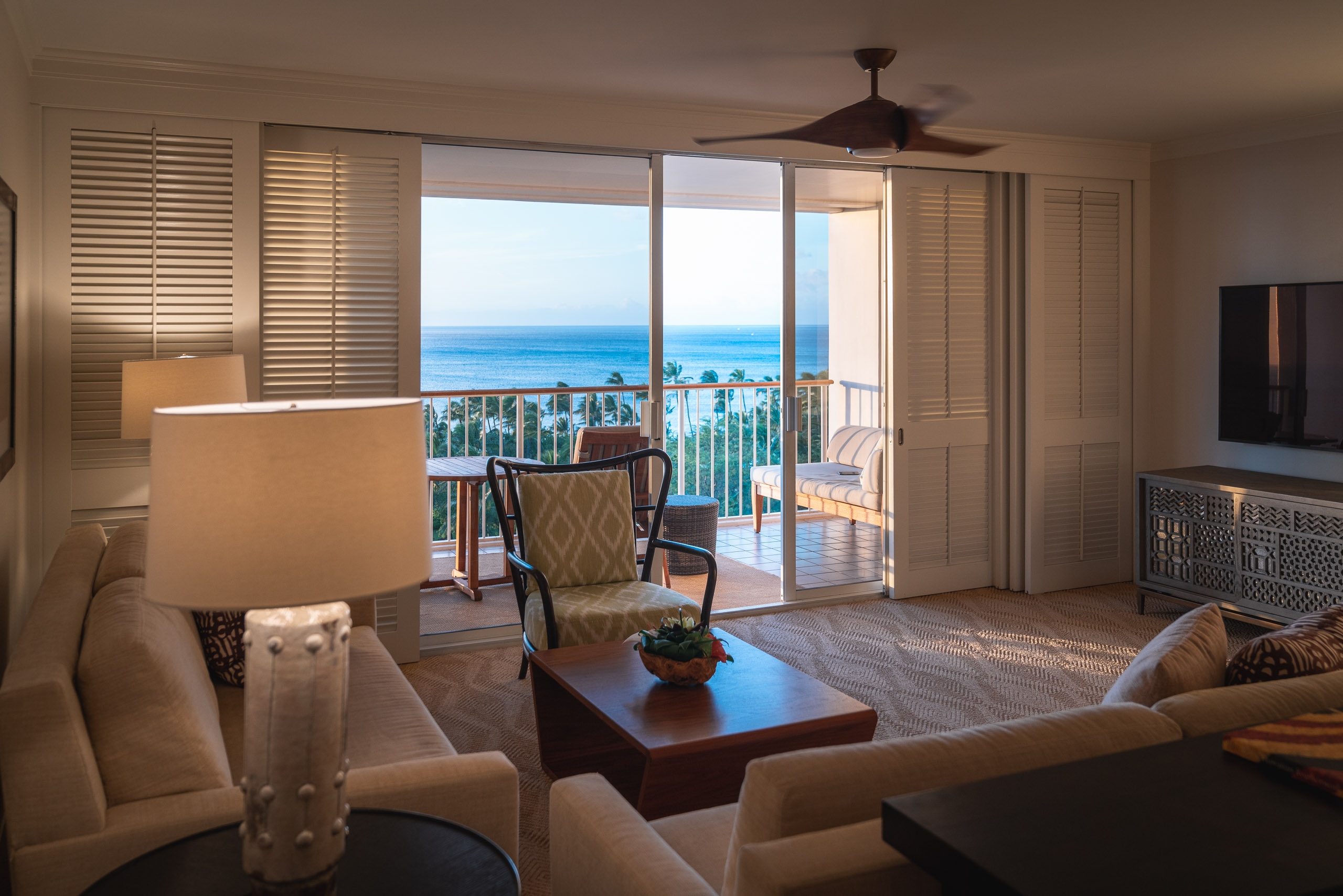 Luxury oceanfront condominium interior with cream-toned furnishings, dark wood accents, and expansive glass doors opening onto a balcony overlooking turquoise Hawaiian waters