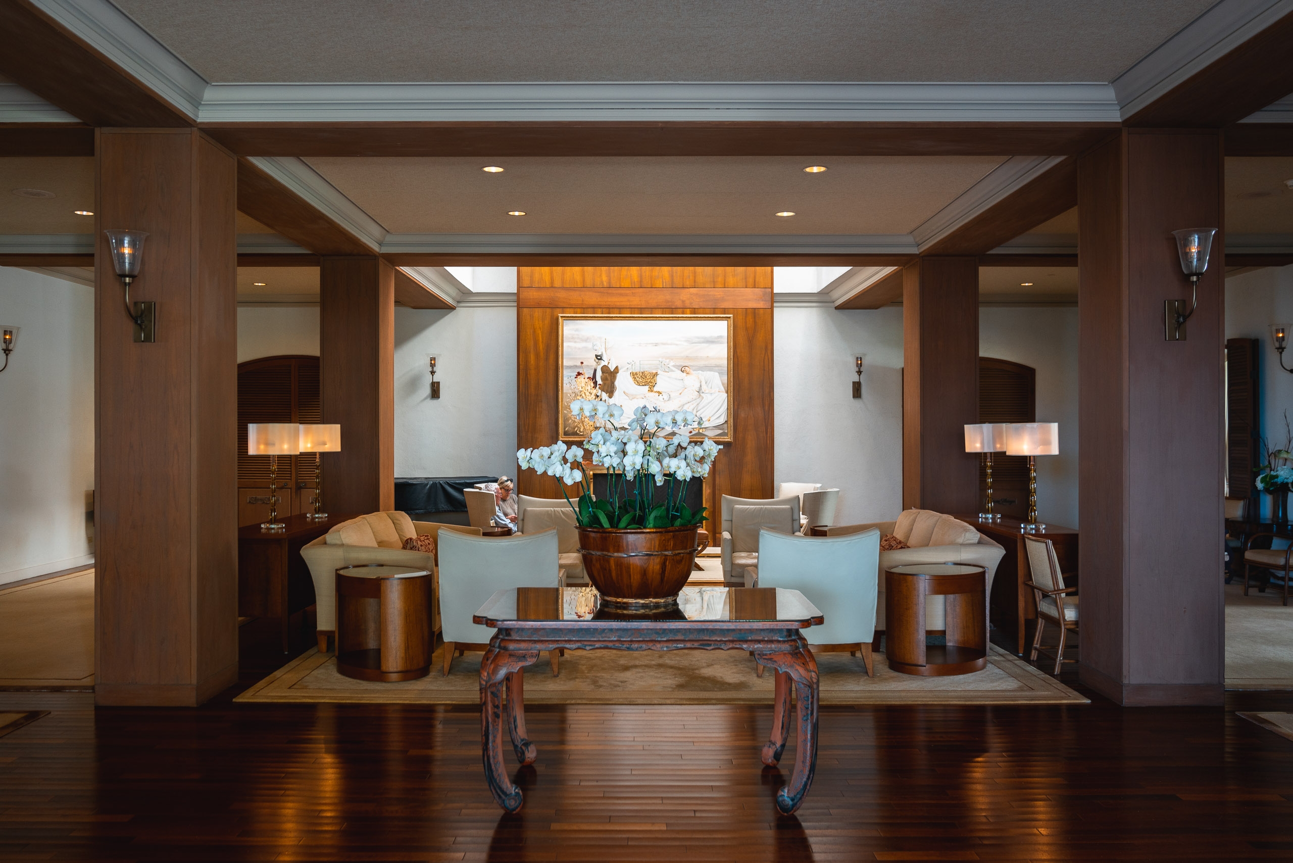 A symmetrical luxury hotel lobby features rich wood paneling, paired light blue armchairs flanking a carved table, and white orchids in a copper vessel, with warm recessed lighting and ocean views framed by the central doorway