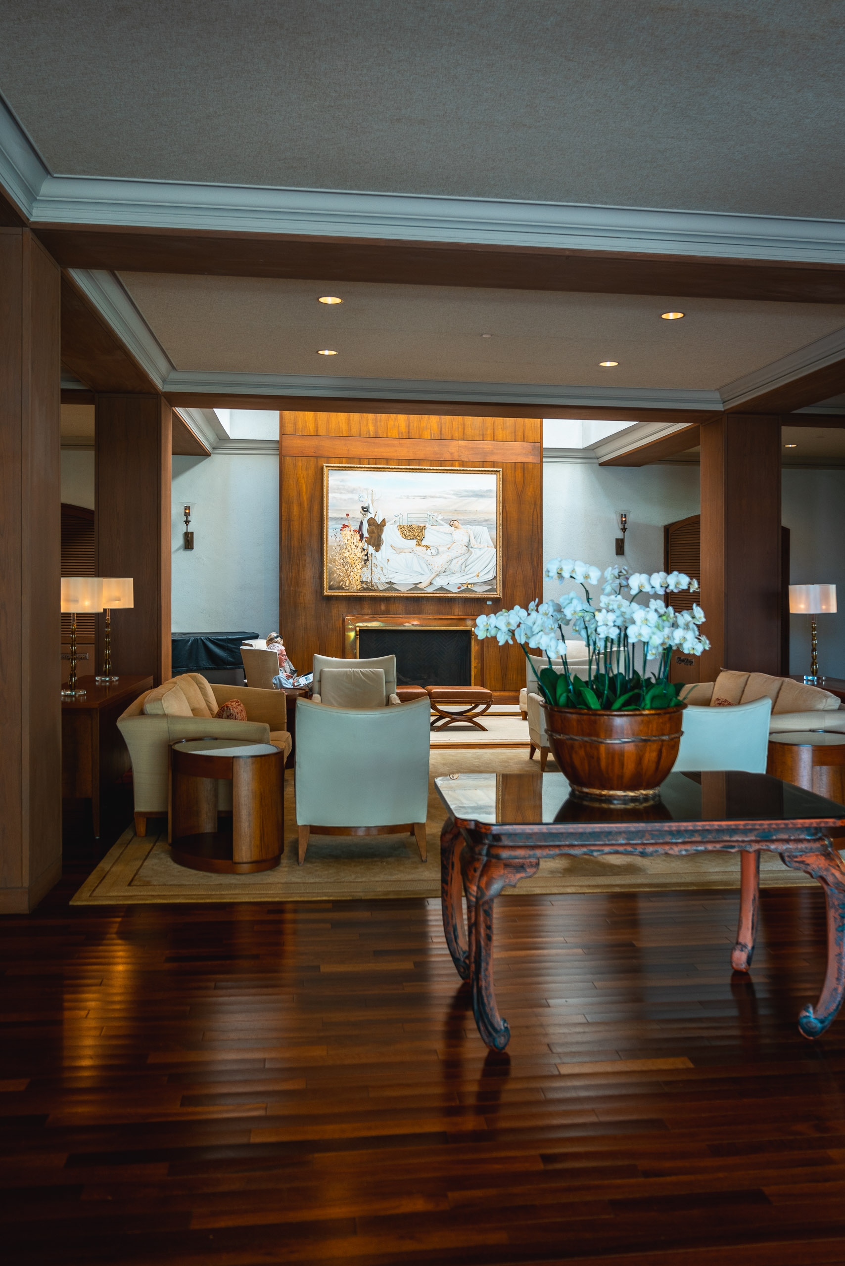 A modern luxury hotel suite features rich wood flooring, light blue ceiling details, and an arrangement of pale blue upholstered chairs beside a decorative wooden table holding white orchids in a bronze vessel