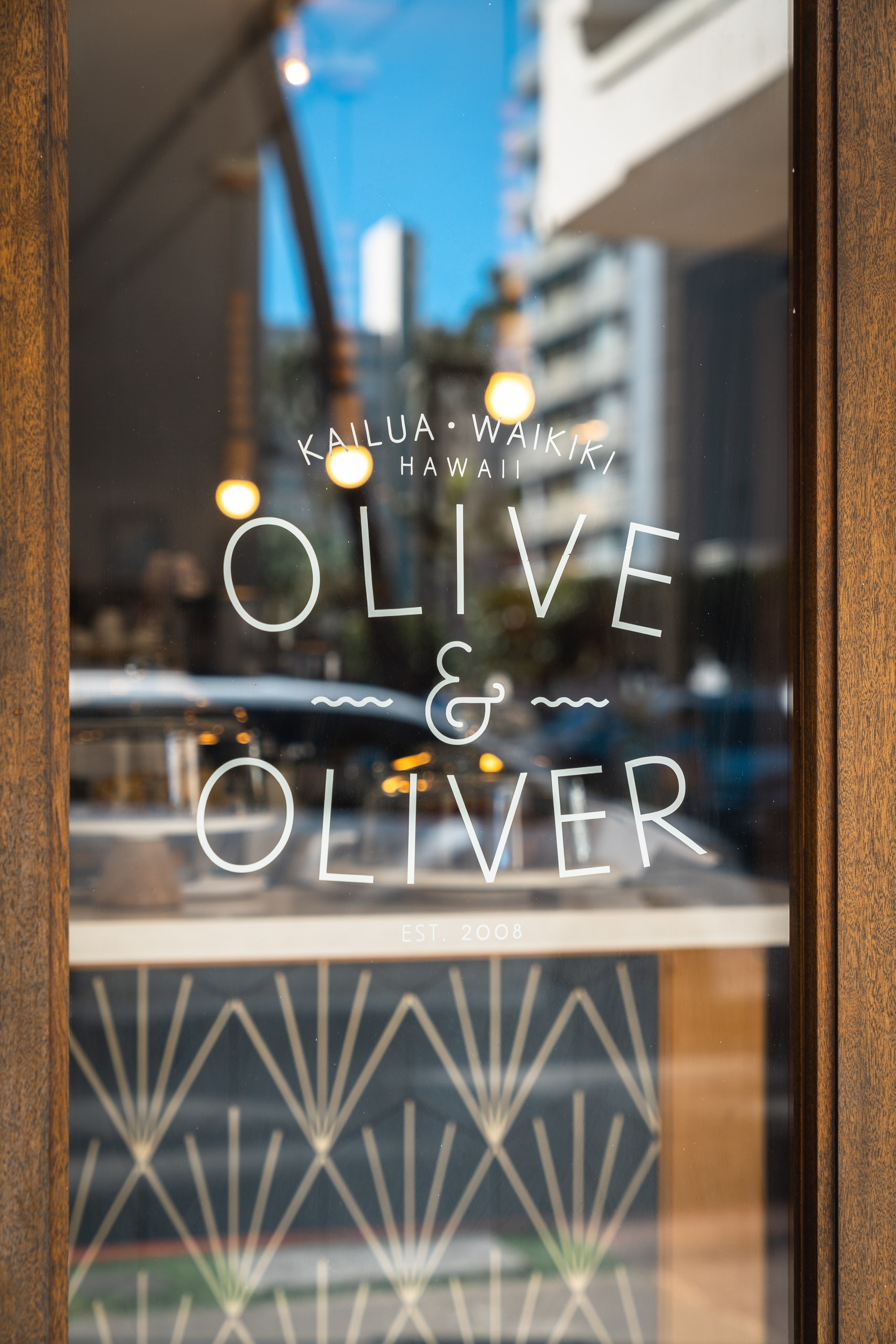 Restaurant storefront window in Waikiki displays white neon lettering reading "Olive & Oliver" with warm pendant lights glowing above, reflecting the blue Hawaiian sky and modern buildings beyond