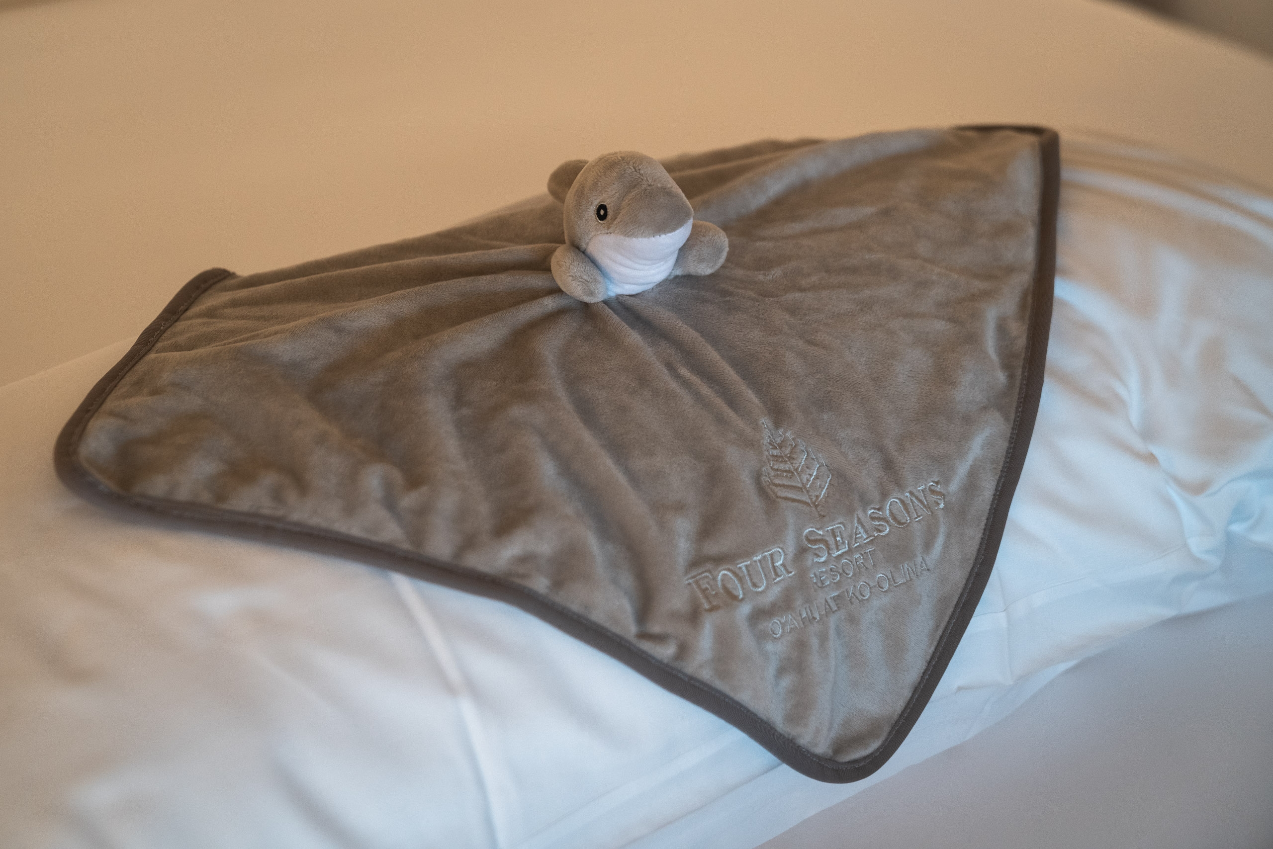 A small gray plush toy with a white mask sits atop a folded Four Seasons branded blanket on white bedding, captured in soft indoor lighting
