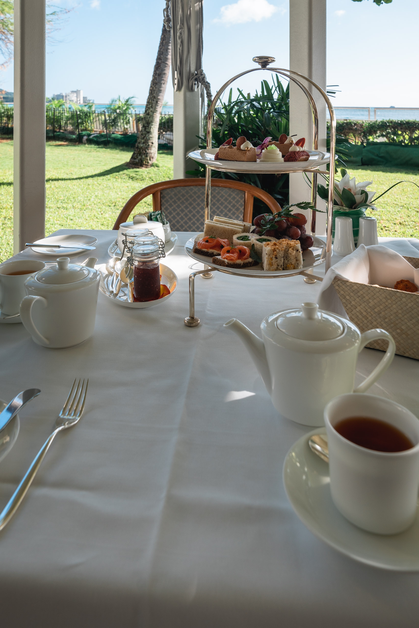 An elegant afternoon tea service set on a white-draped table overlooks manicured tropical gardens and distant ocean, with tiered pastries, white porcelain teapots, and fresh fruit arranged against bright Hawaiian daylight