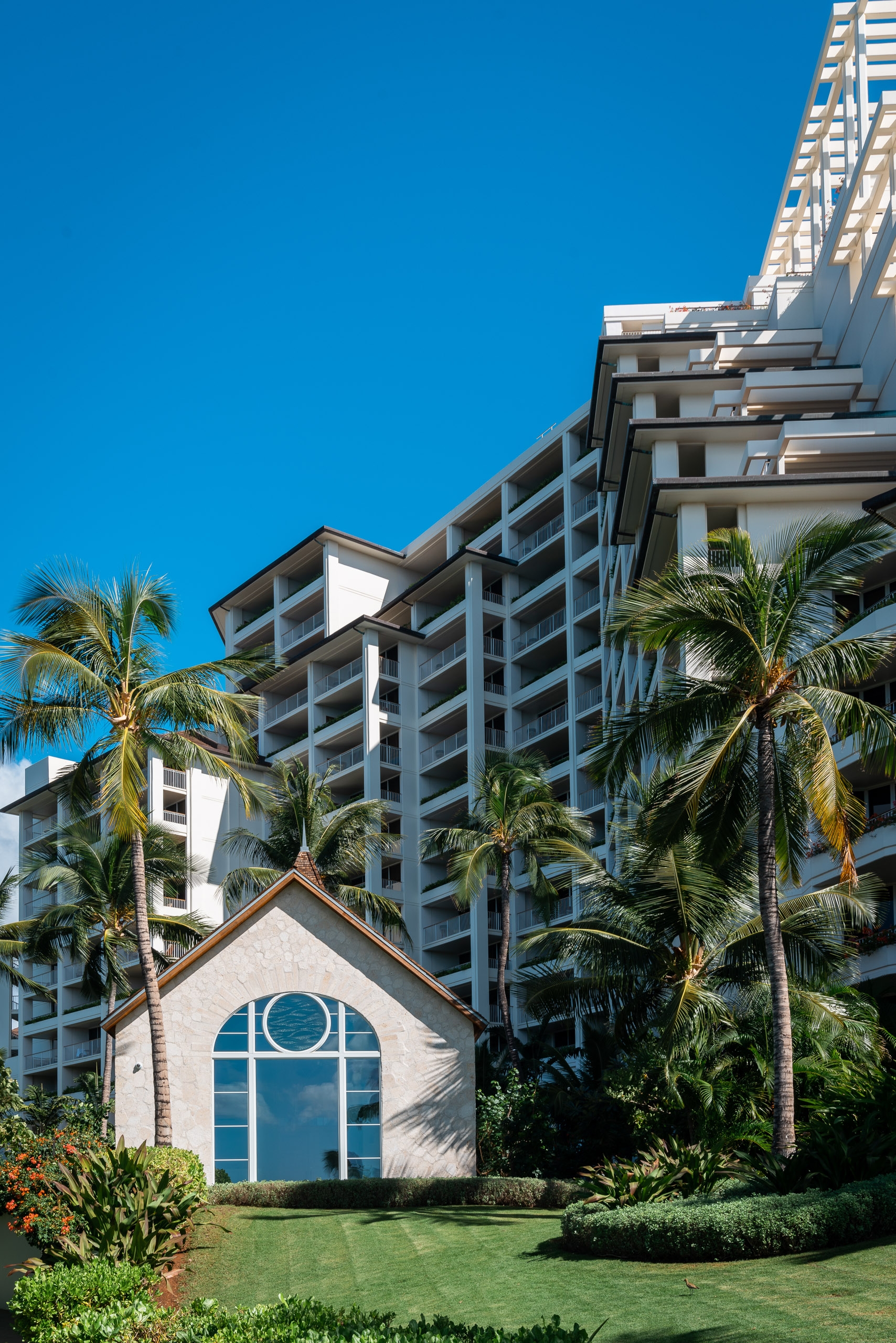Modern beachfront resort buildings tower behind a small white chapel with a circular blue window, flanked by tropical palm trees under clear blue sky