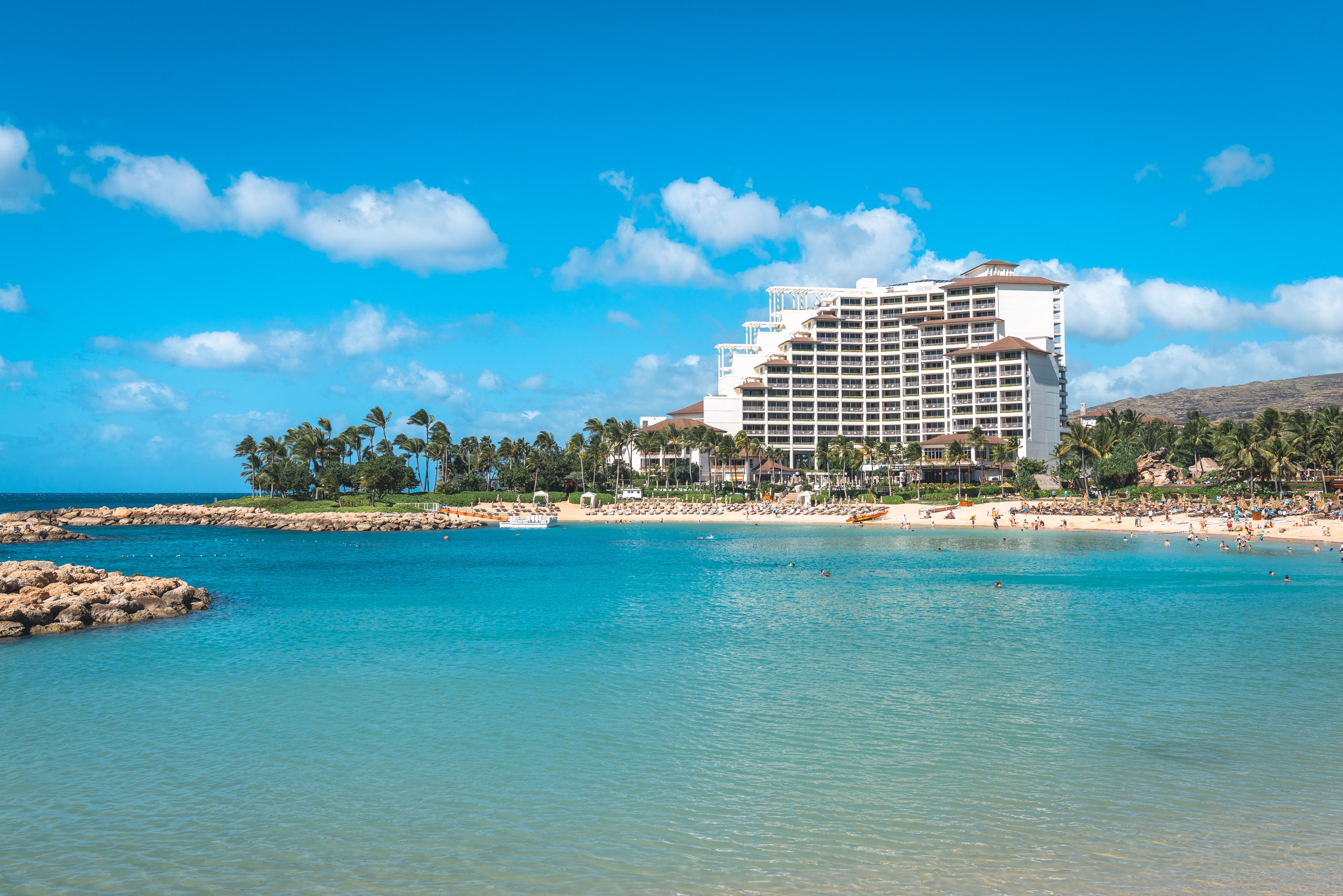 A multi-story beachfront resort sits beside a crescent cove with turquoise water, palm trees, and sandy beach under clear blue sky with white clouds