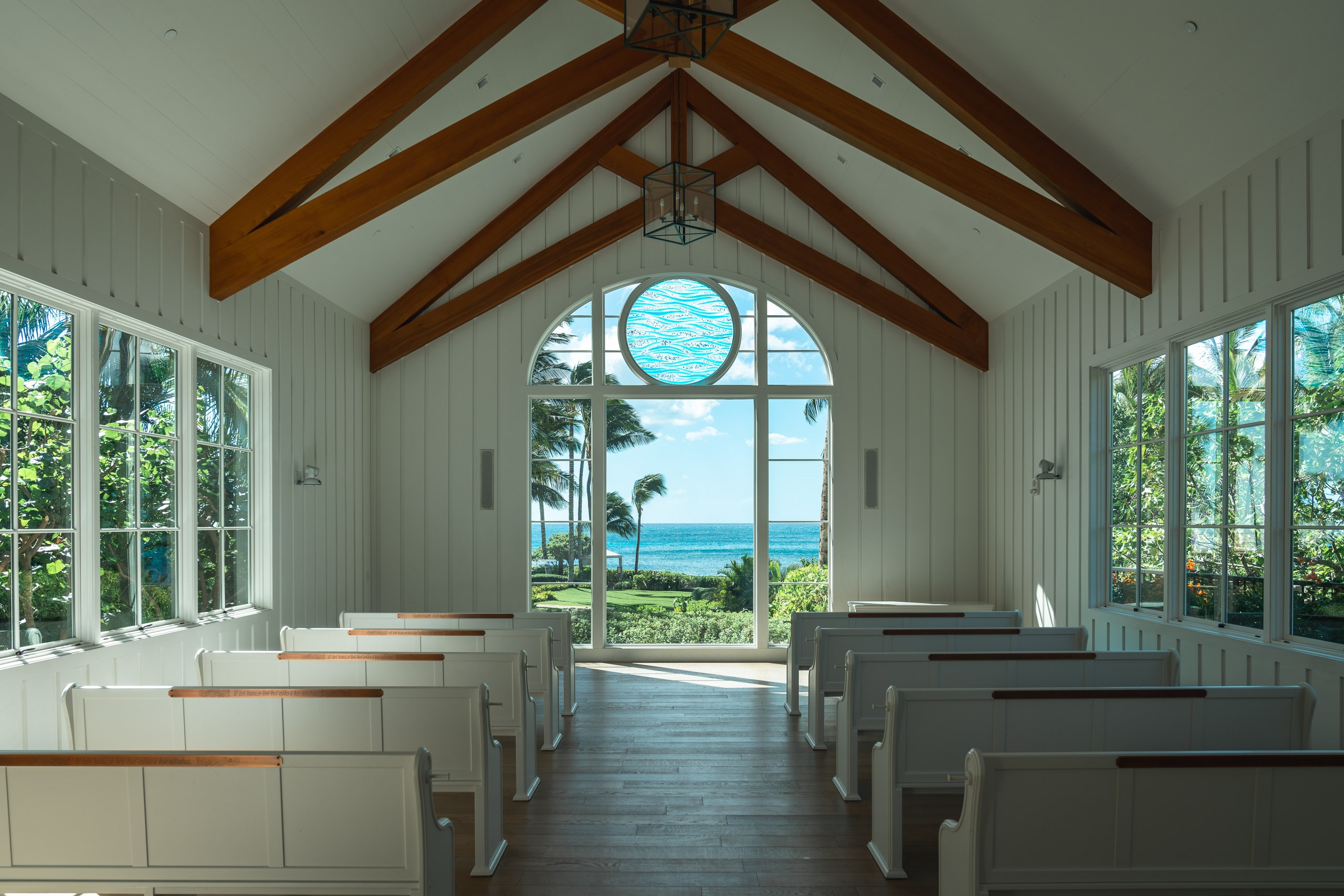 A bright chapel interior features wooden ceiling beams, white pews lining both sides, and expansive windows framing an ocean view with palm trees and blue sky beyond