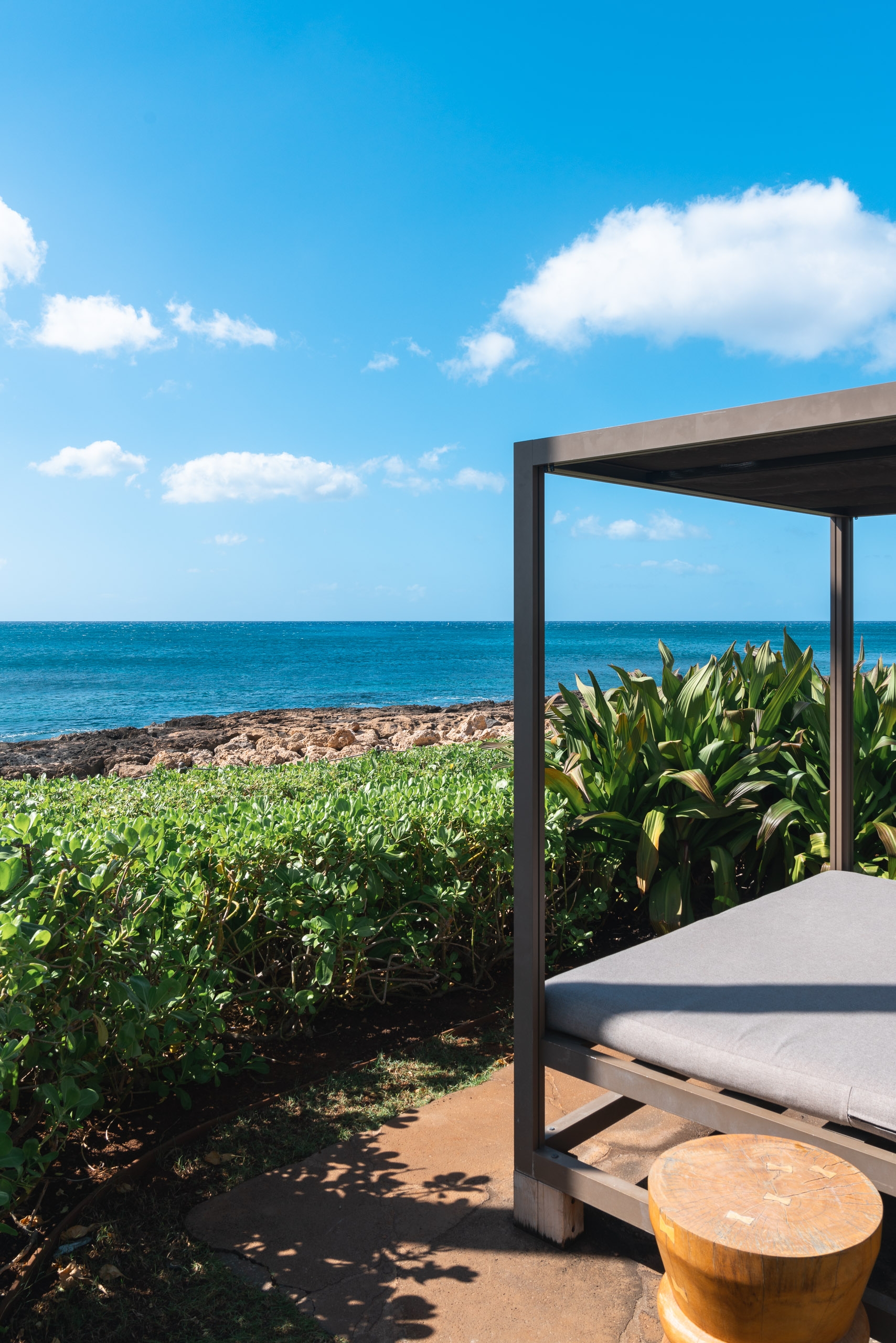 A modern cabana with white cushioned daybed overlooks a turquoise ocean and rocky shoreline, with lush green vegetation and white clouds dotting the bright blue sky