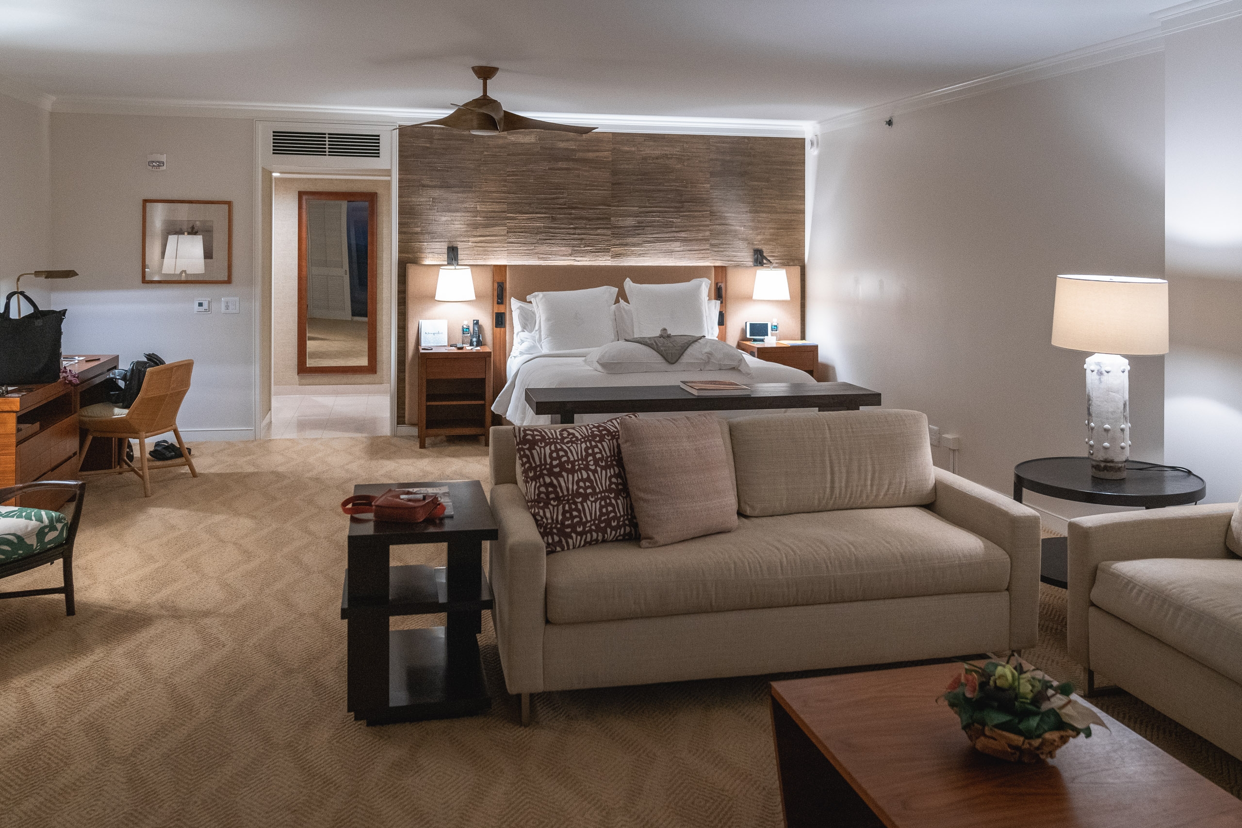 Spacious luxury hotel suite featuring a beige sofa and seating area in the foreground, with a wooden-headboard bed, warm lighting, and neutral tones throughout the well-appointed room