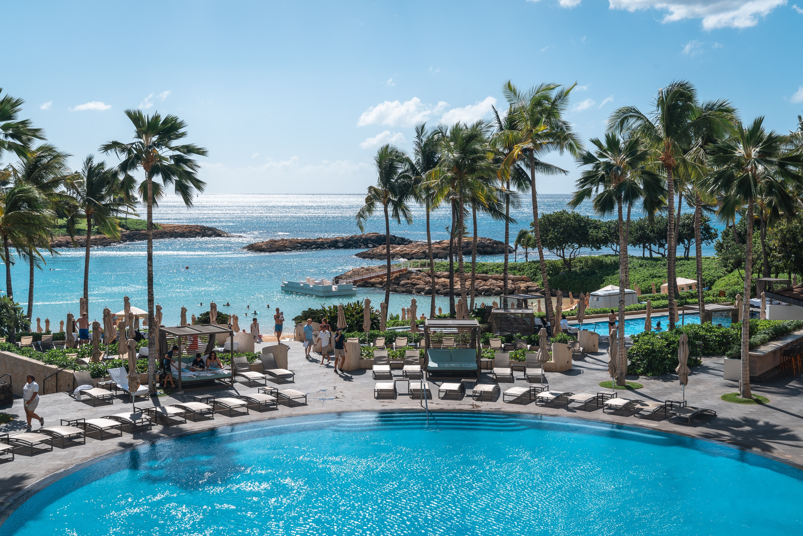 A luxury resort pool with turquoise water sits surrounded by palm trees, lounge chairs, and guests, overlooking a calm turquoise cove and ocean horizon under blue sky