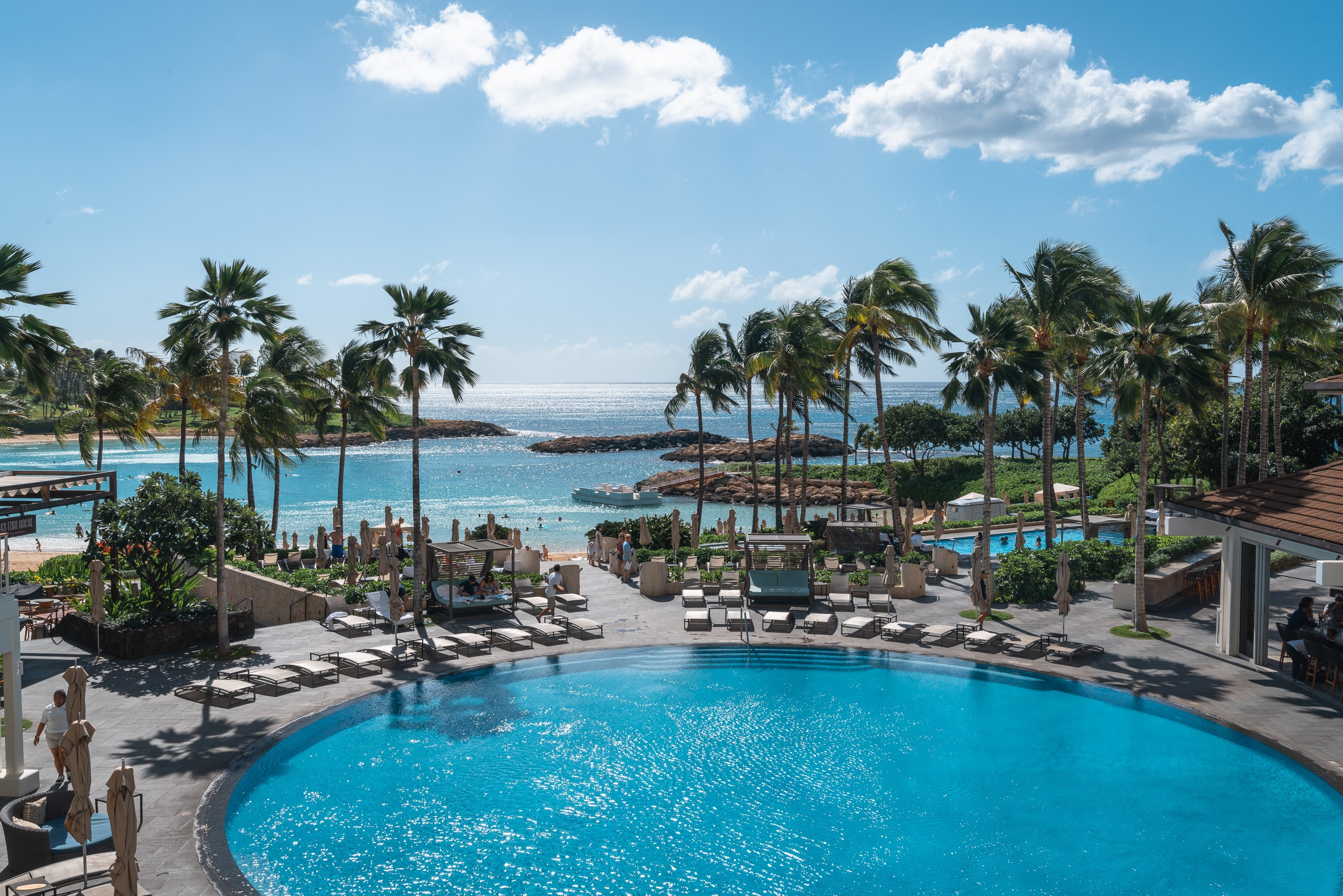A luxurious resort pool overlooks a turquoise lagoon fringed with palm trees, white sand beach, and calm ocean waters under bright blue sky with scattered clouds