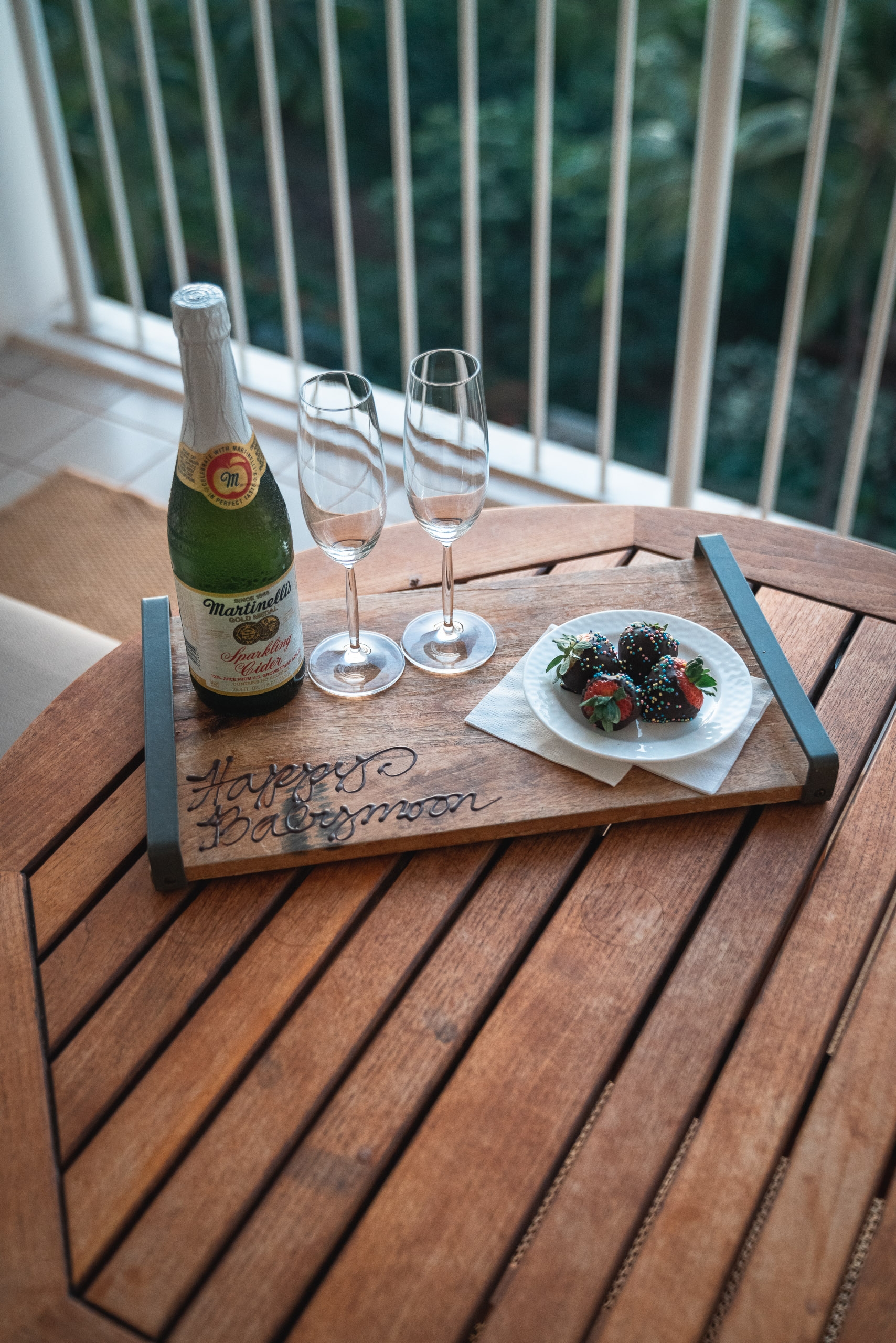 A wooden deck table holds a bottle of Matilda Bay sparkling wine, two empty glasses, and a small plate of berries with greenery, with a railed balcony and lush foliage visible beyond