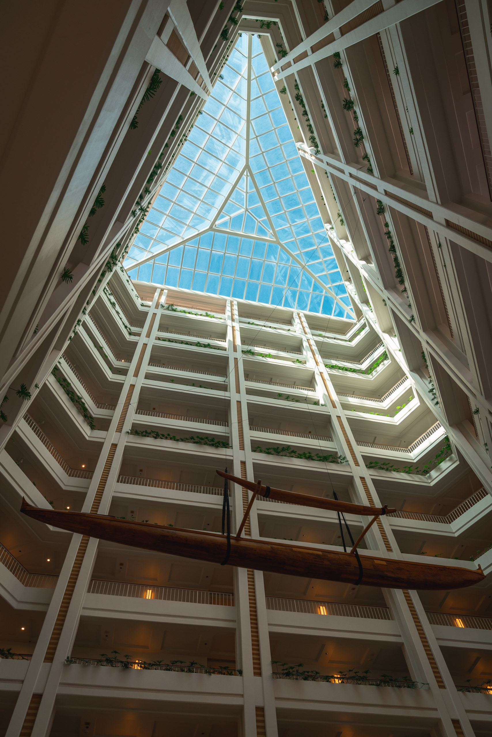 A dramatic upward view of a modern hotel atrium reveals multiple tiered balconies with white railings converging toward a geometric glass ceiling framing bright blue sky above