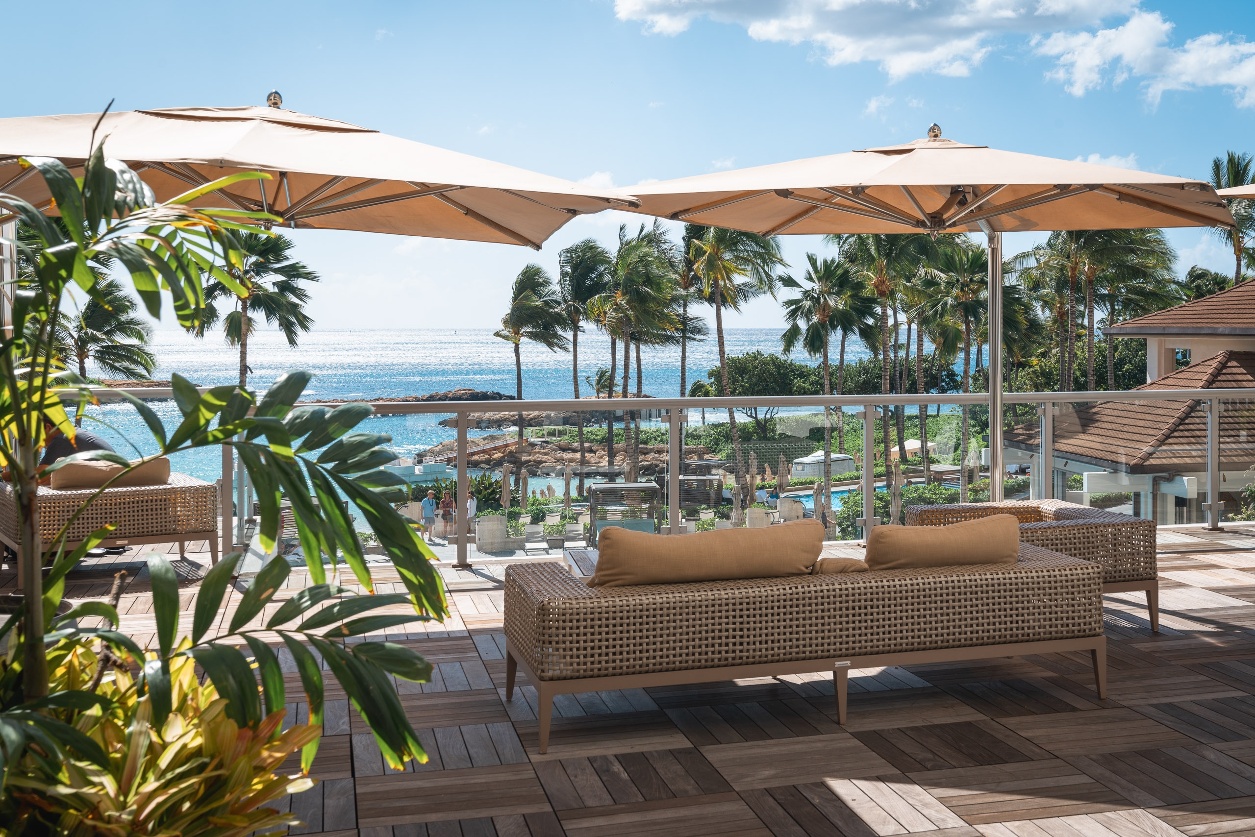 A modern resort terrace features woven lounge seating beneath tan umbrellas, overlooking a coastal landscape with palm trees and sparkling ocean waters under blue sky