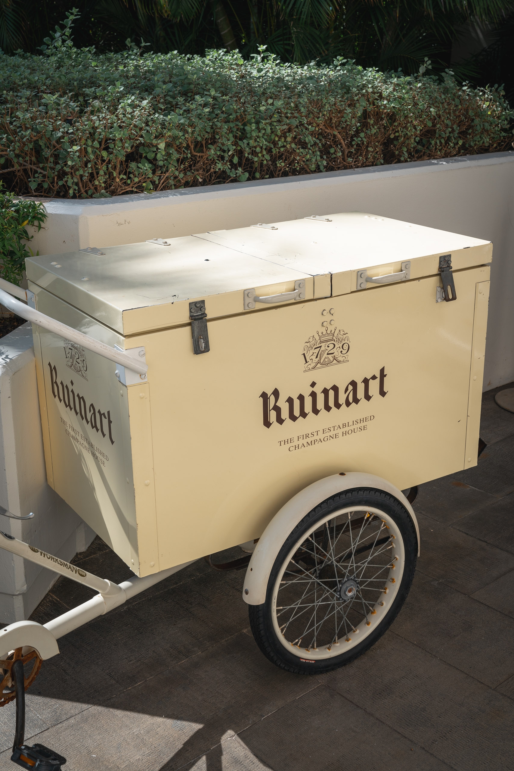 A cream-colored Ruinart champagne promotional cart with white-wall wheels sits on a paved terrace, backed by lush green foliage and dark palm fronds in bright daylight