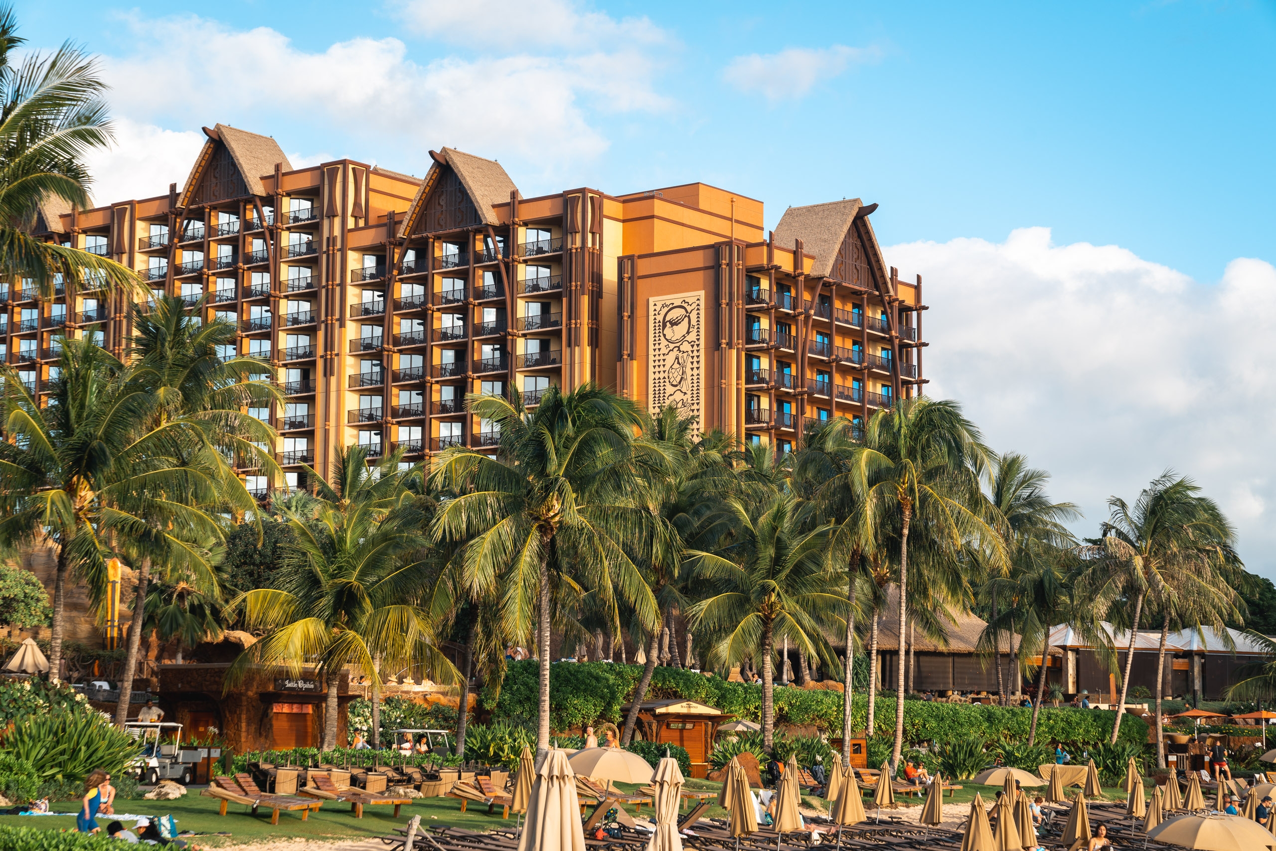 Golden-hued hotel towers with steep rooflines rise behind palm trees and a sandy beach dotted with tan umbrellas and loungers under clear blue sky