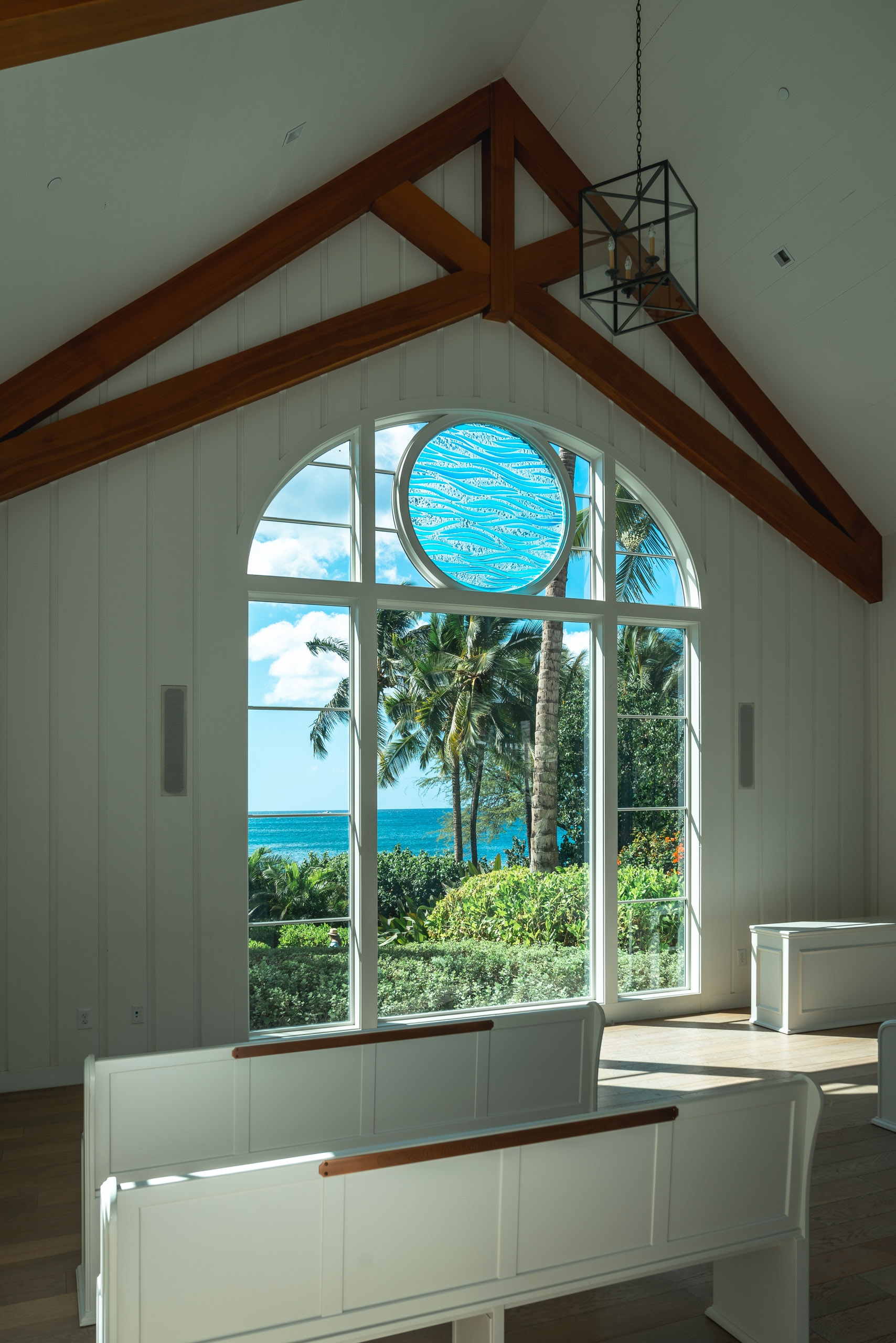 Bright interior with exposed wooden beams frames a large arched window overlooking tropical palms and turquoise ocean, bathed in natural sunlight.