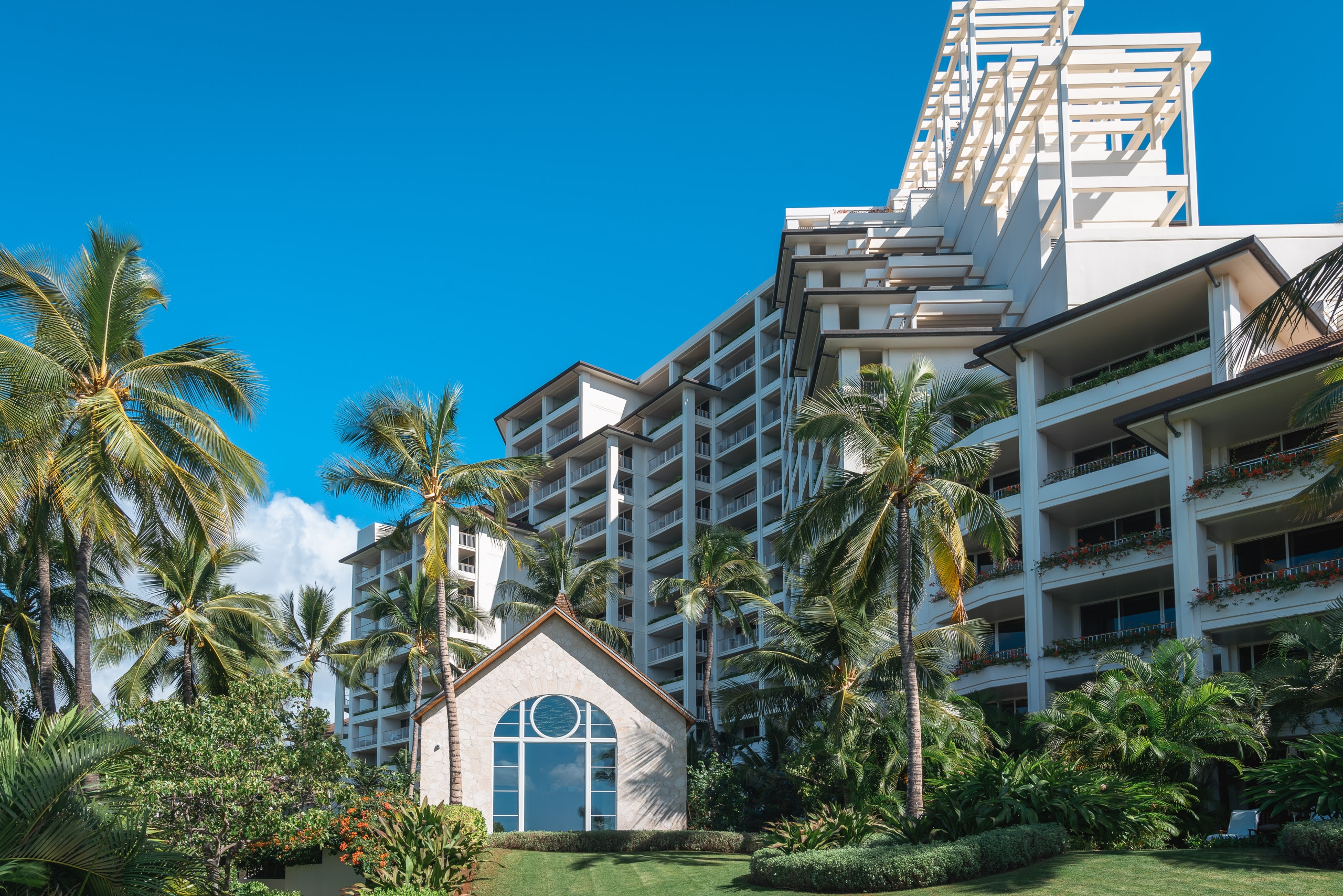 Modern white resort buildings with multiple balconies rise behind a small chapel structure, surrounded by swaying palm trees under bright blue sky in a tropical Hawaiian setting