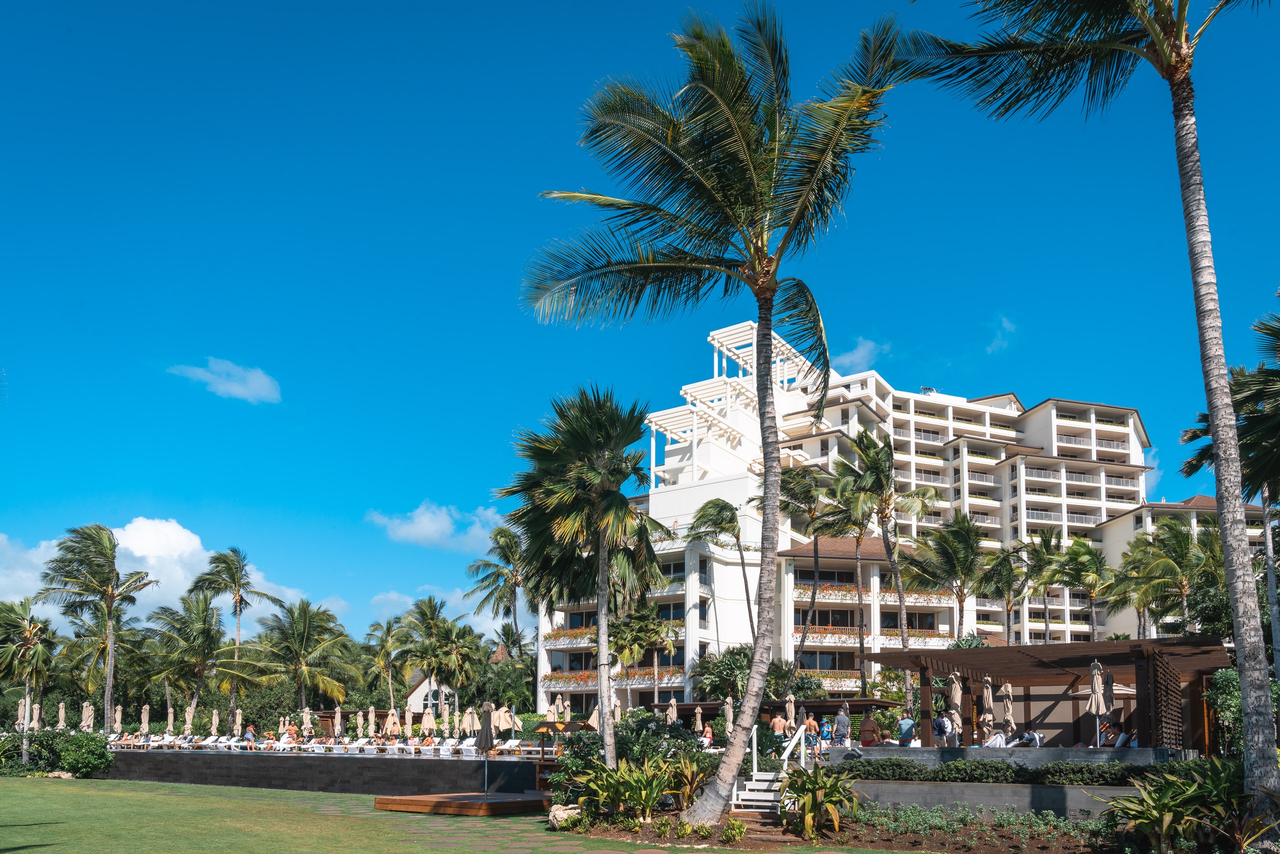 A multi-story white resort hotel stands amid tall palm trees under clear blue sky, with manicured green lawn and beachgoers visible in the foreground at this Hawaiian coastal property
