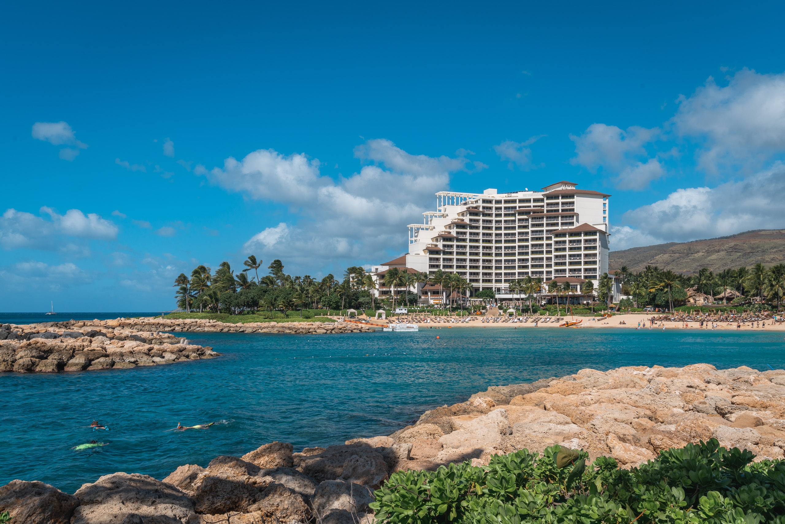 A large beachfront resort hotel stands on a sandy shore lined with palm trees, viewed across turquoise waters dotted with swimmers and framed by rocky outcroppings under a bright blue sky with white clouds