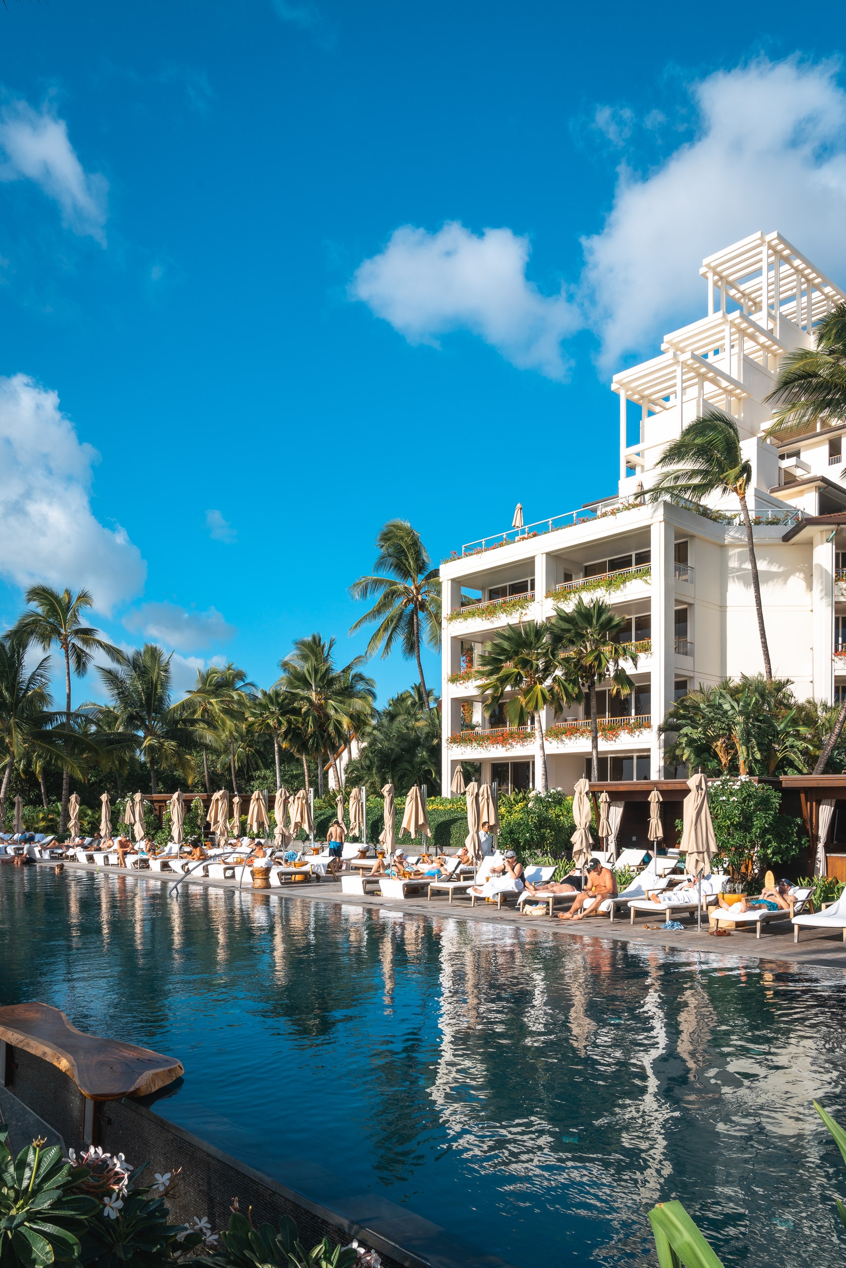 A white multi-story resort building with palm trees stands beside a calm lagoon lined with beige umbrellas and lounge chairs under bright blue skies with scattered white clouds