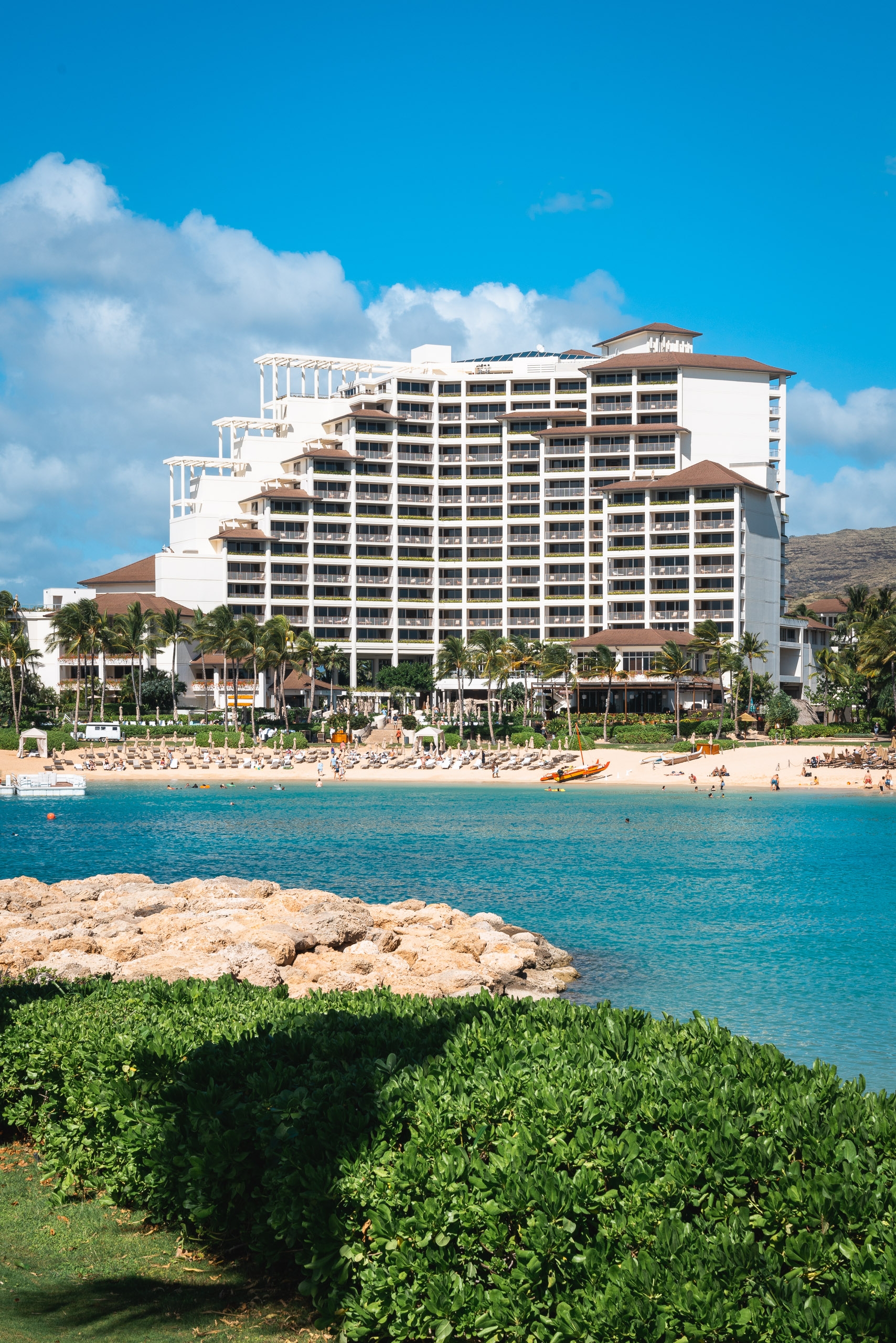 A beachfront resort hotel rises above turquoise waters and sandy shores, with palm trees and green vegetation framing the scene under a bright blue sky with white clouds