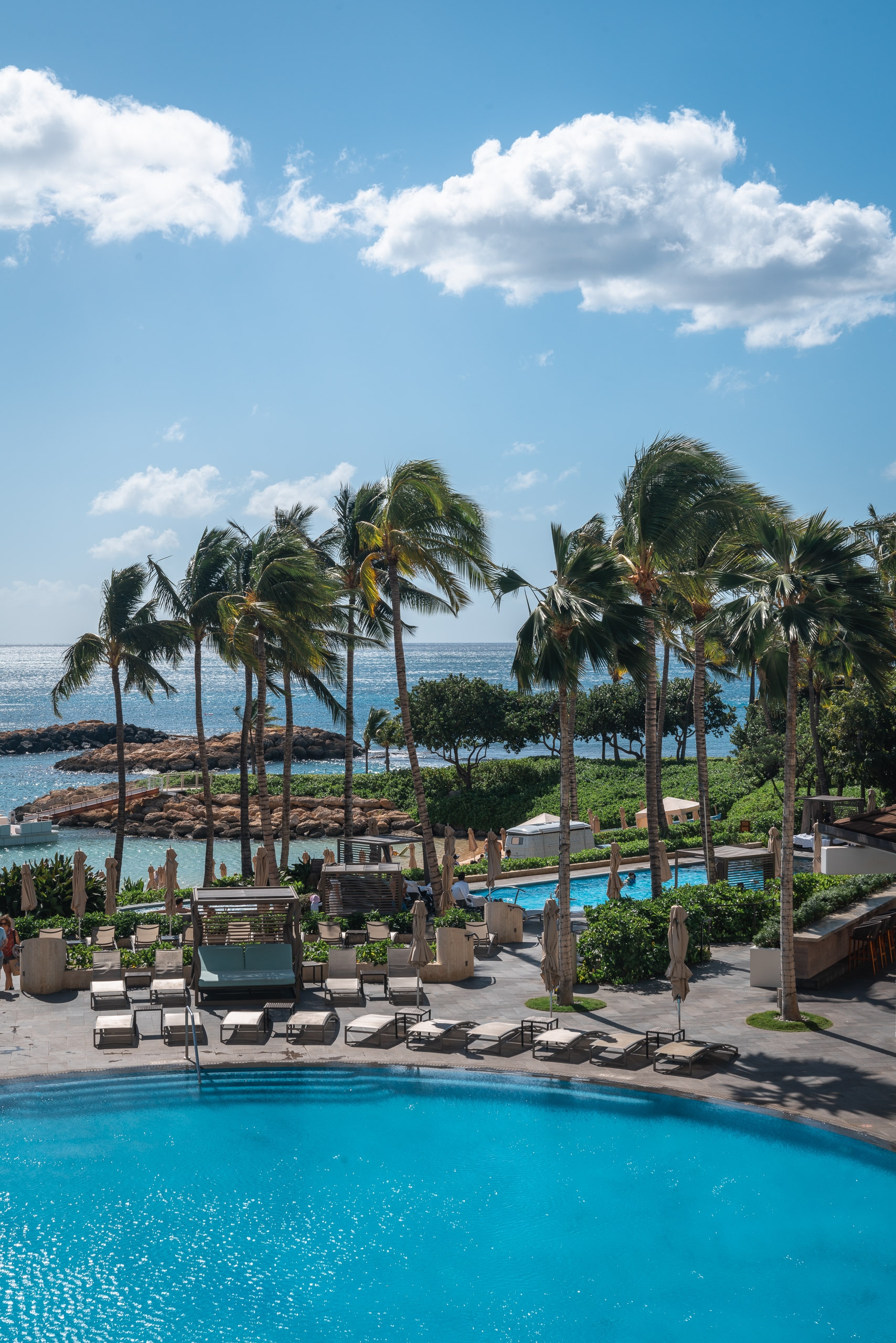 A luxurious resort pool with bright turquoise water overlooks palm-lined grounds, ocean vistas, and white clouds against a clear blue sky in Hawaii