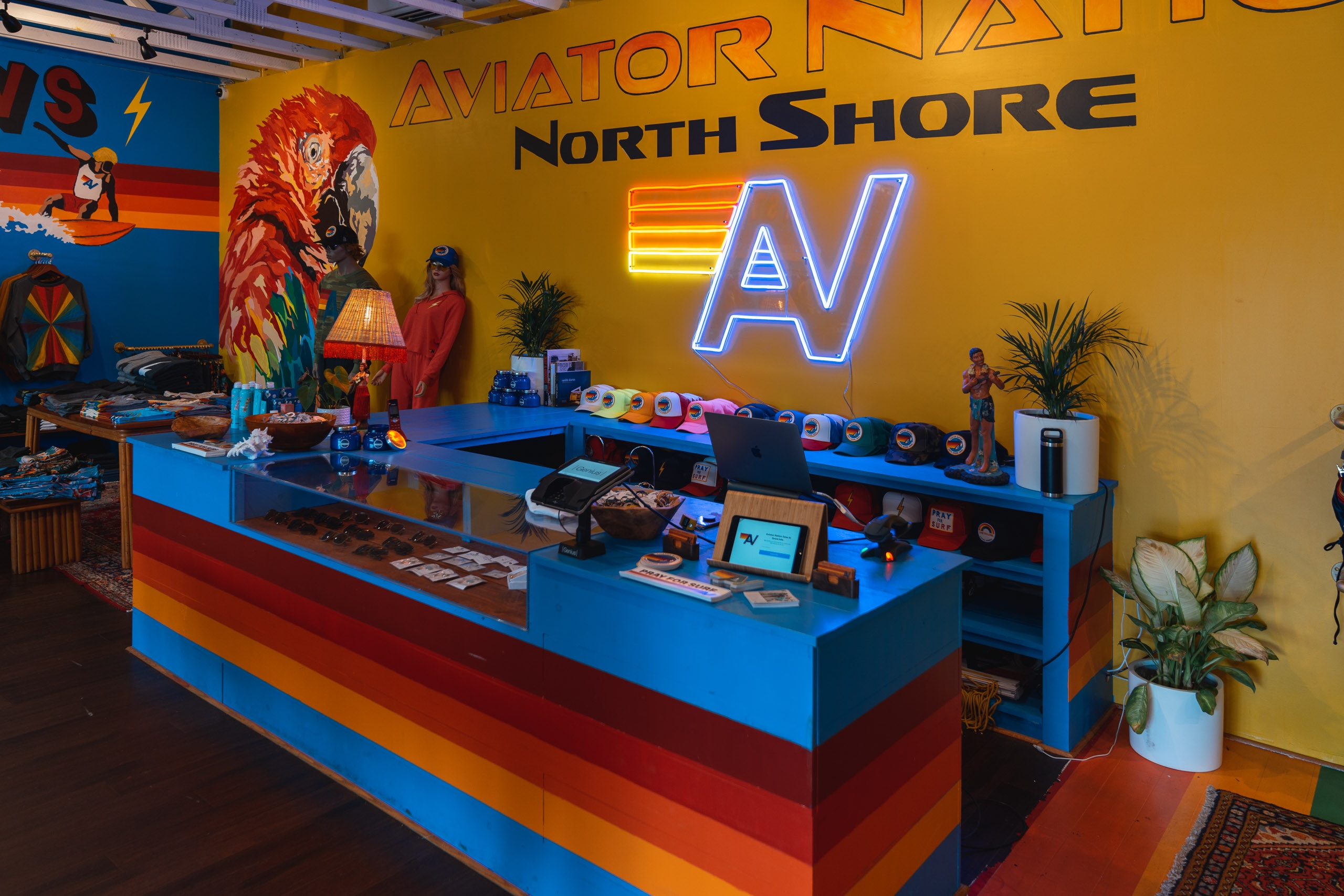 A vibrant retail counter with blue, red, and orange striped front displays merchandise under bright neon signage reading "Aviator North Shore" against a bold yellow wall decorated with colorful aviation-themed artwork and plants