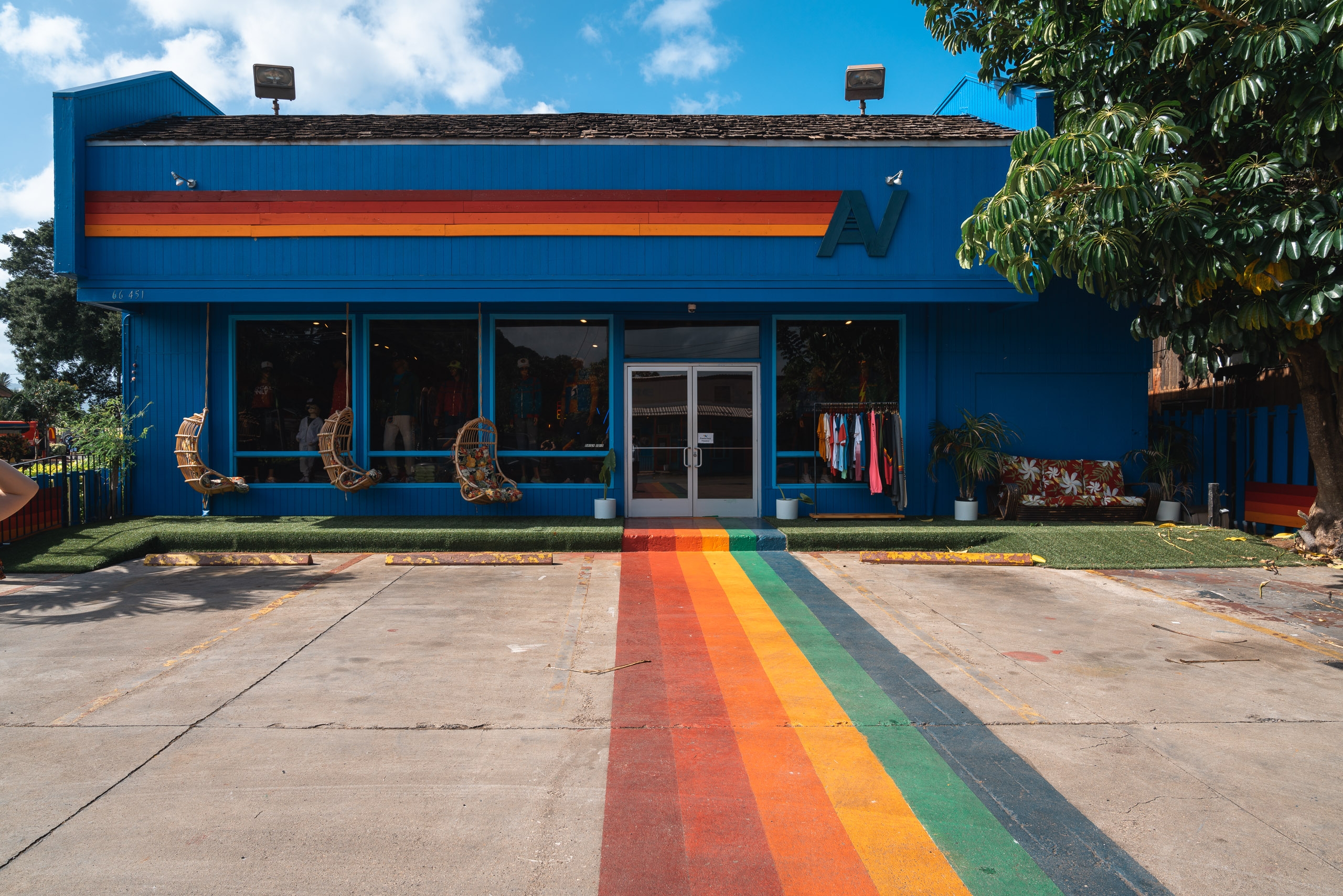 A vibrant blue building with an orange stripe features a rainbow-painted walkway leading to its entrance, set against clear skies and tropical foliage