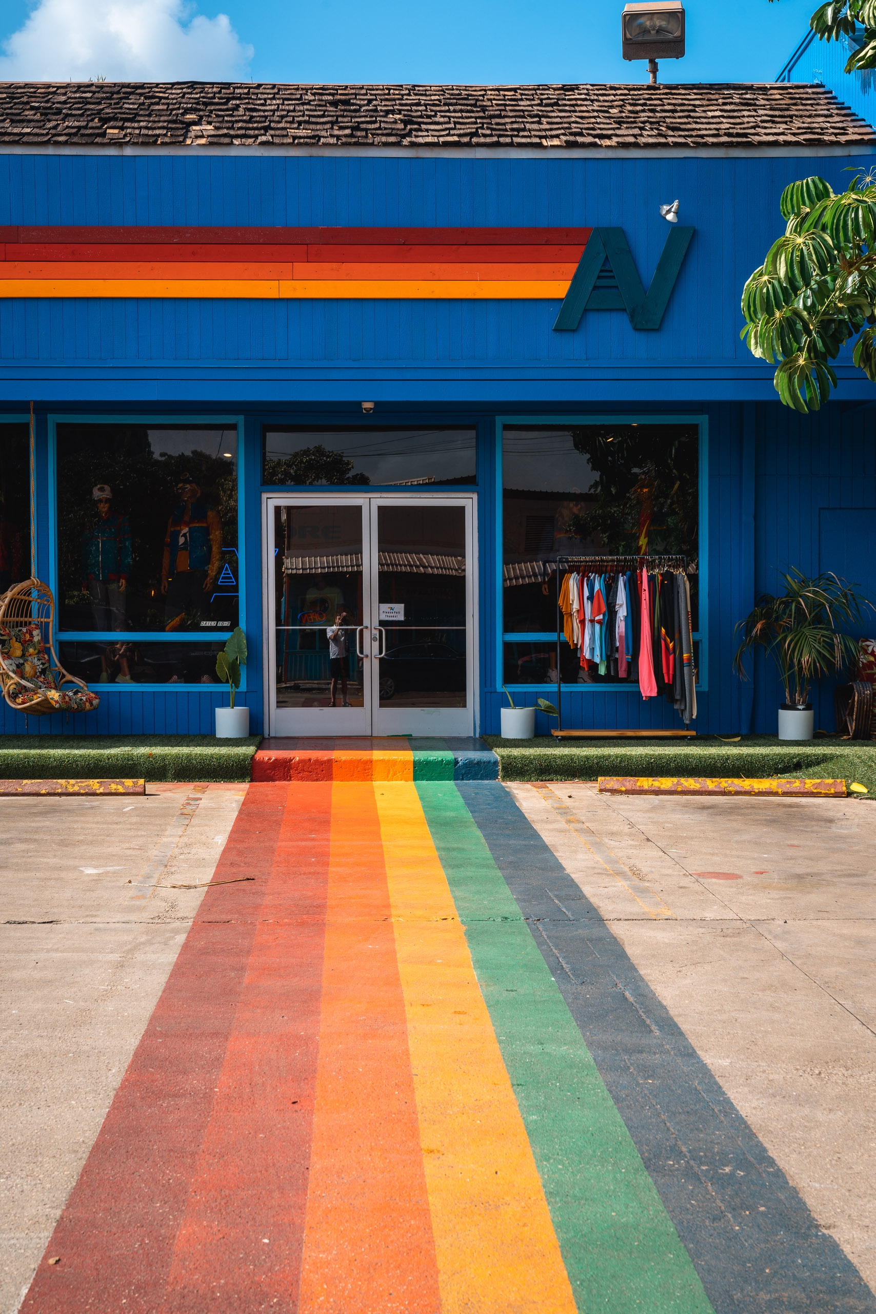 A bright blue storefront with rainbow-striped pavement leading to its entrance displays clothing racks visible through large windows under clear skies