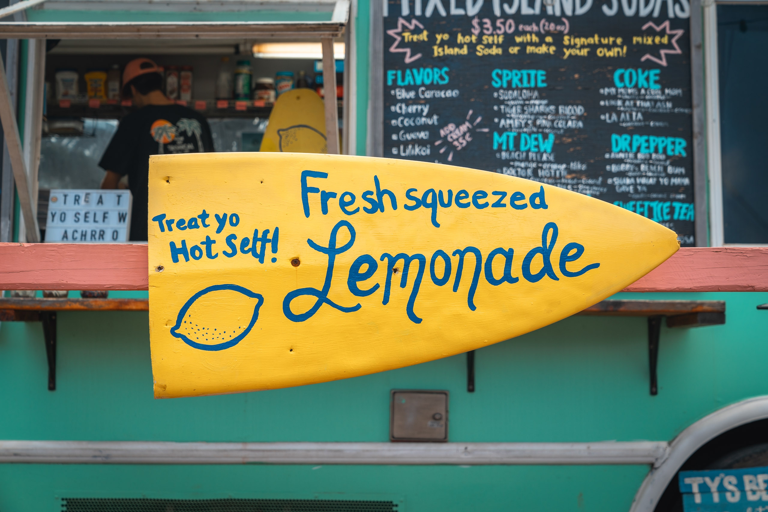 A bright yellow lemon-shaped sign advertising fresh squeezed lemonade hangs on a turquoise food truck at a casual beachside vendor stand