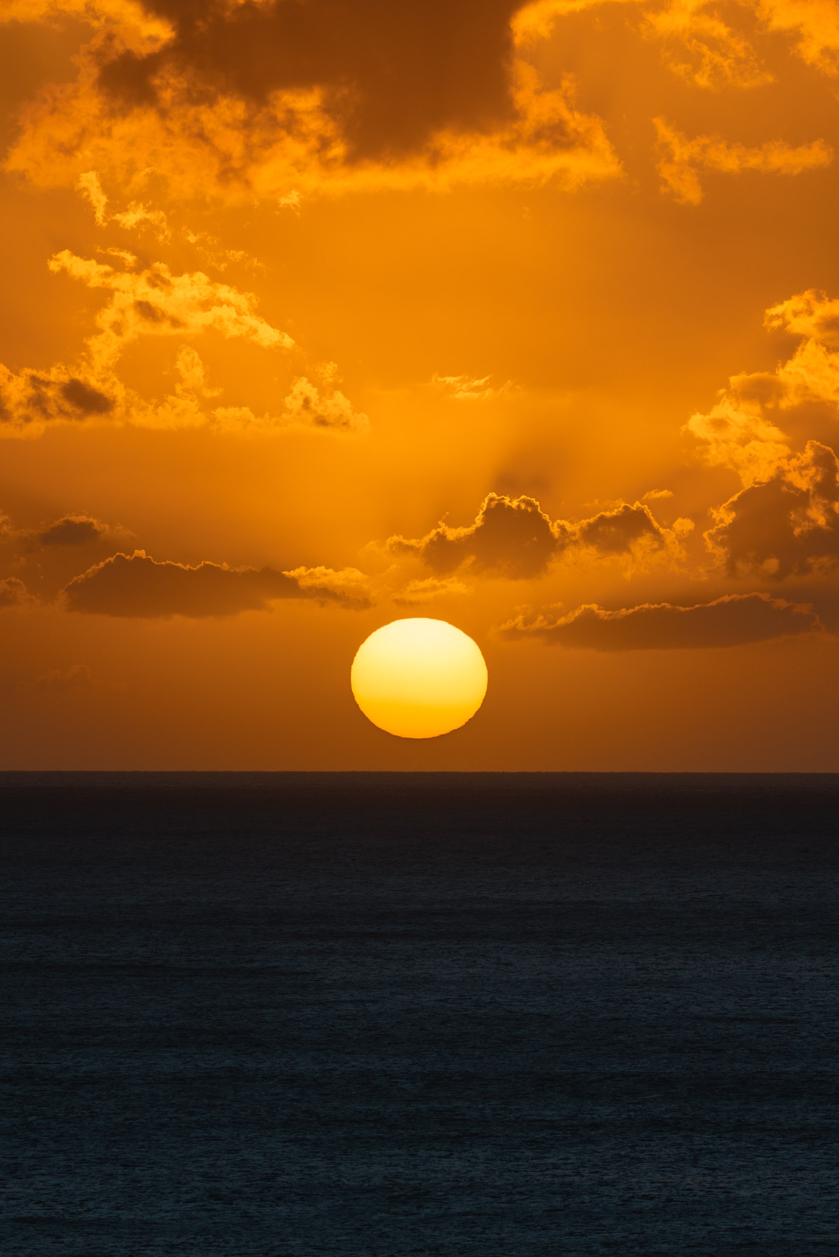 A golden sun rests on the ocean horizon beneath an amber sky filled with scattered dark clouds at dusk