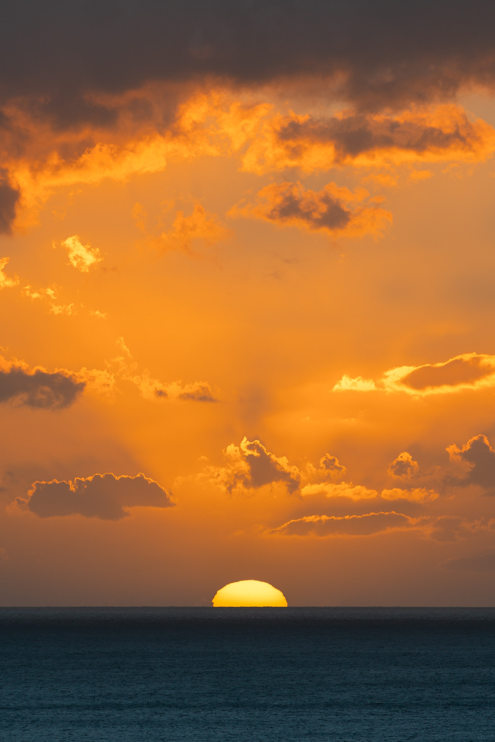 The sun's golden disk touches the horizon over calm ocean waters while dramatic orange and amber clouds fill the sky above during a serene sunset