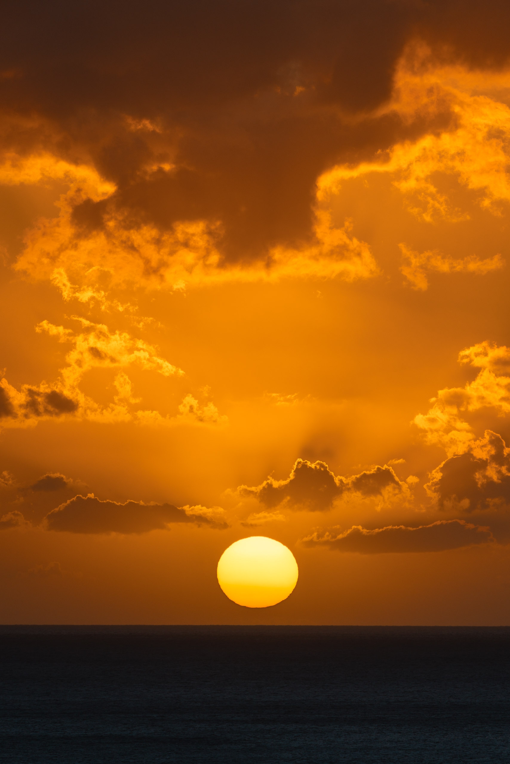 The sun descends toward a calm ocean horizon, casting warm golden light across dramatic clouds tinged with orange and brown tones at dusk