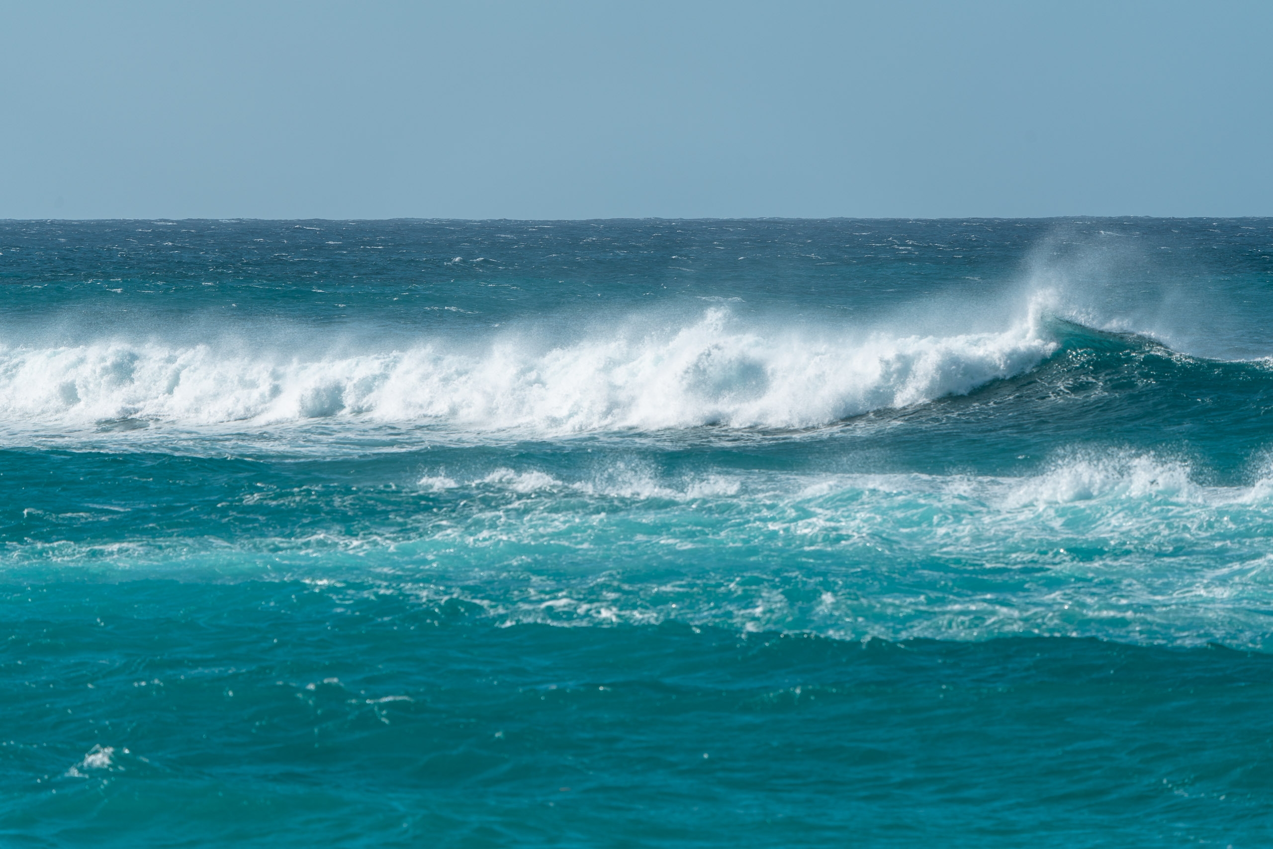 Layered ocean waves with white foam crash across turquoise and deep blue waters under a clear pale sky, capturing the powerful surf conditions of Hawaii's North Shore