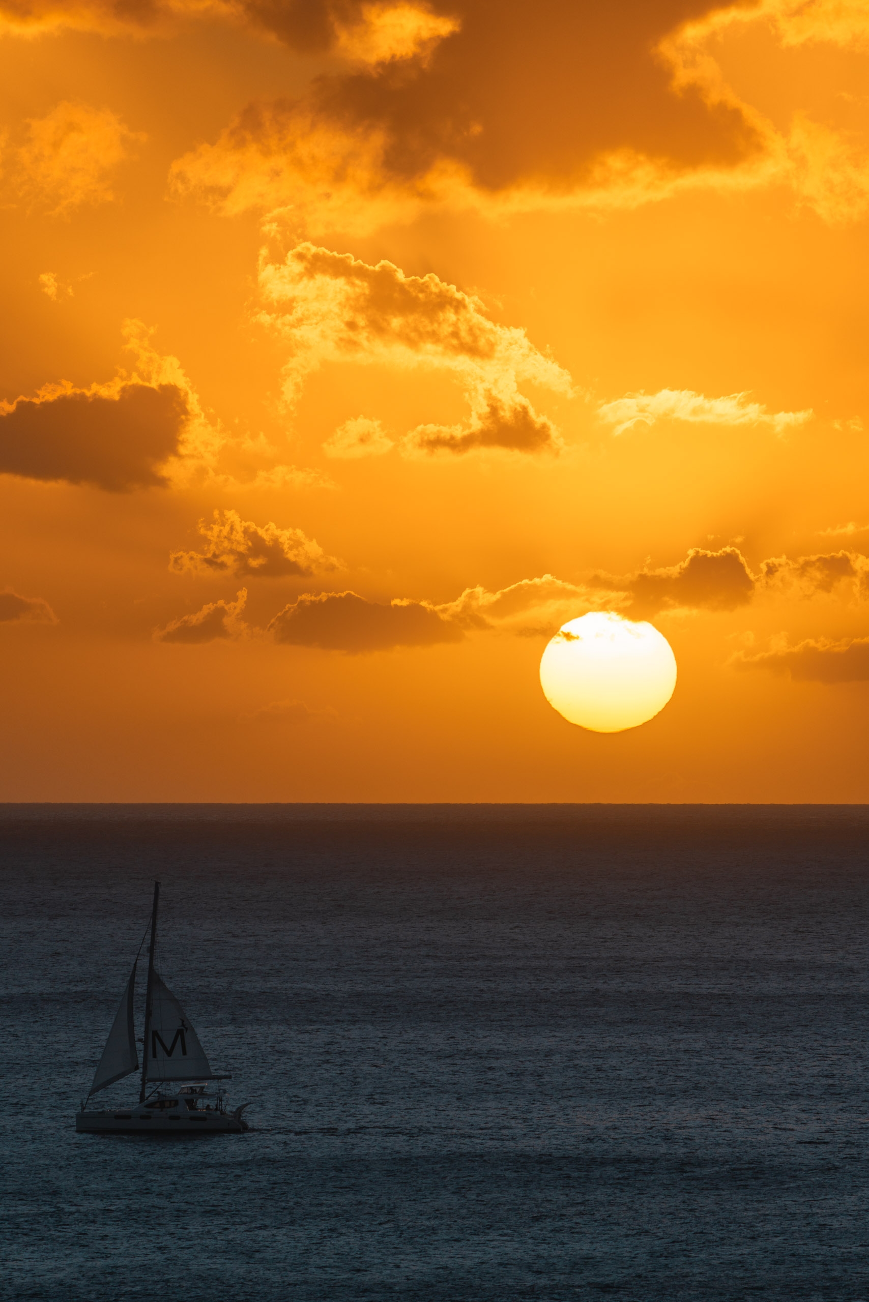 A sailboat silhouetted against calm ocean waters floats beneath a golden sunset sky with dramatic cloud formations and a luminous sun hanging near the horizon