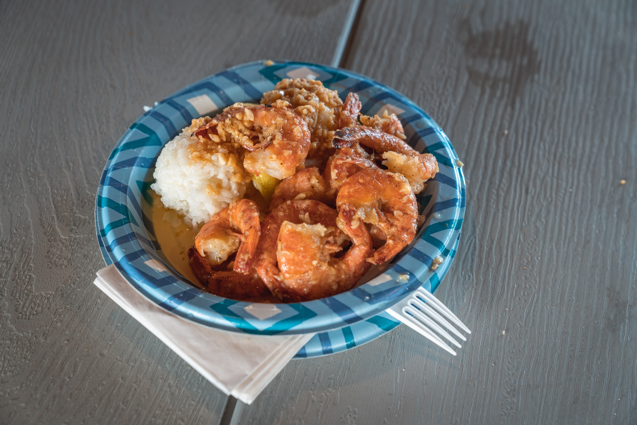 Shrimp scampi served over white rice in a blue-checkered paper bowl with a plastic fork on a wooden table