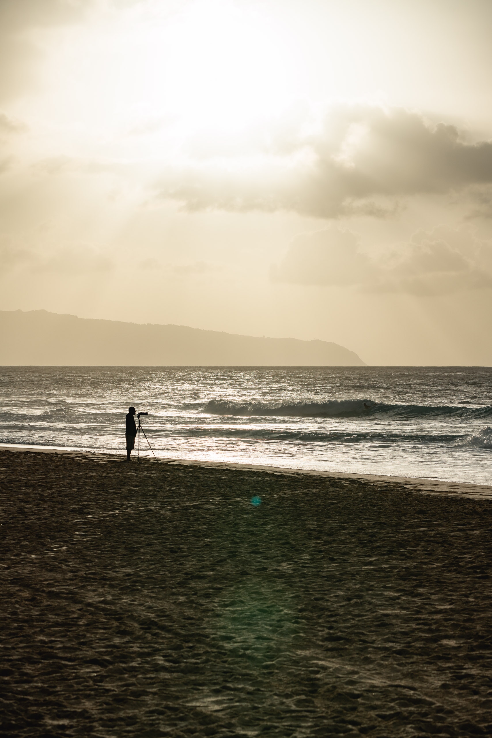 A surfer in a wetsuit stands with a surfboard on a dark sand beach facing rolling ocean waves beneath an overcast sky and distant mountains