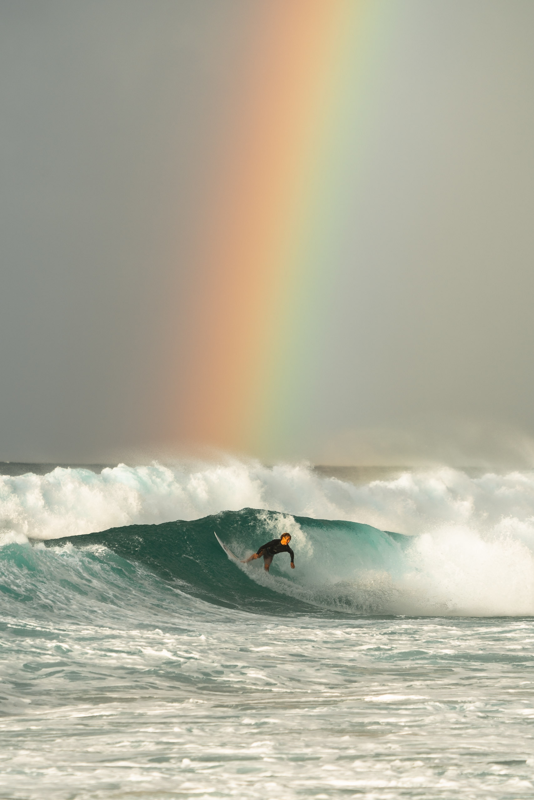 A shirtless surfer rides a turquoise wave beneath a subtle rainbow arcing across an overcast sky and choppy ocean