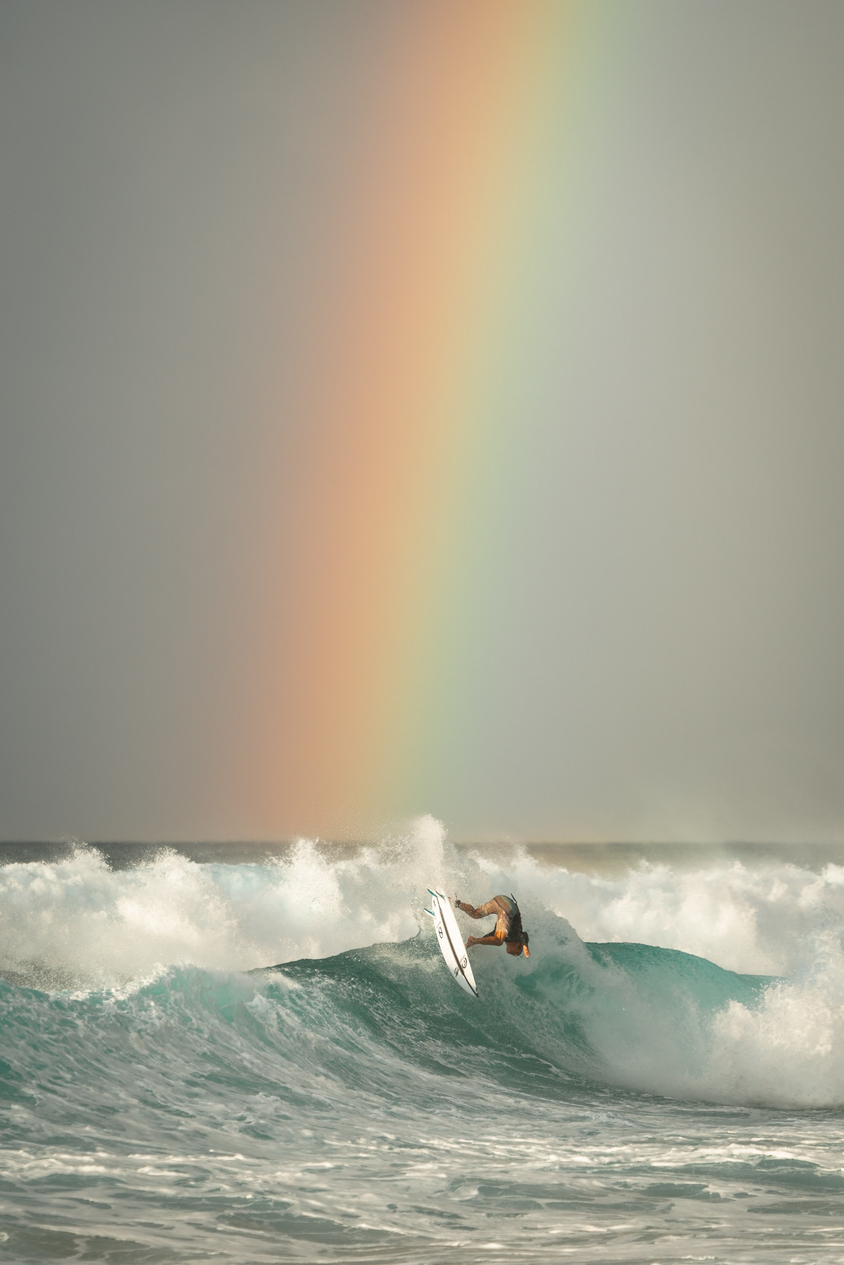 A surfer rides a turquoise wave beneath a delicate rainbow stretching across an overcast sky filled with white spray and foam