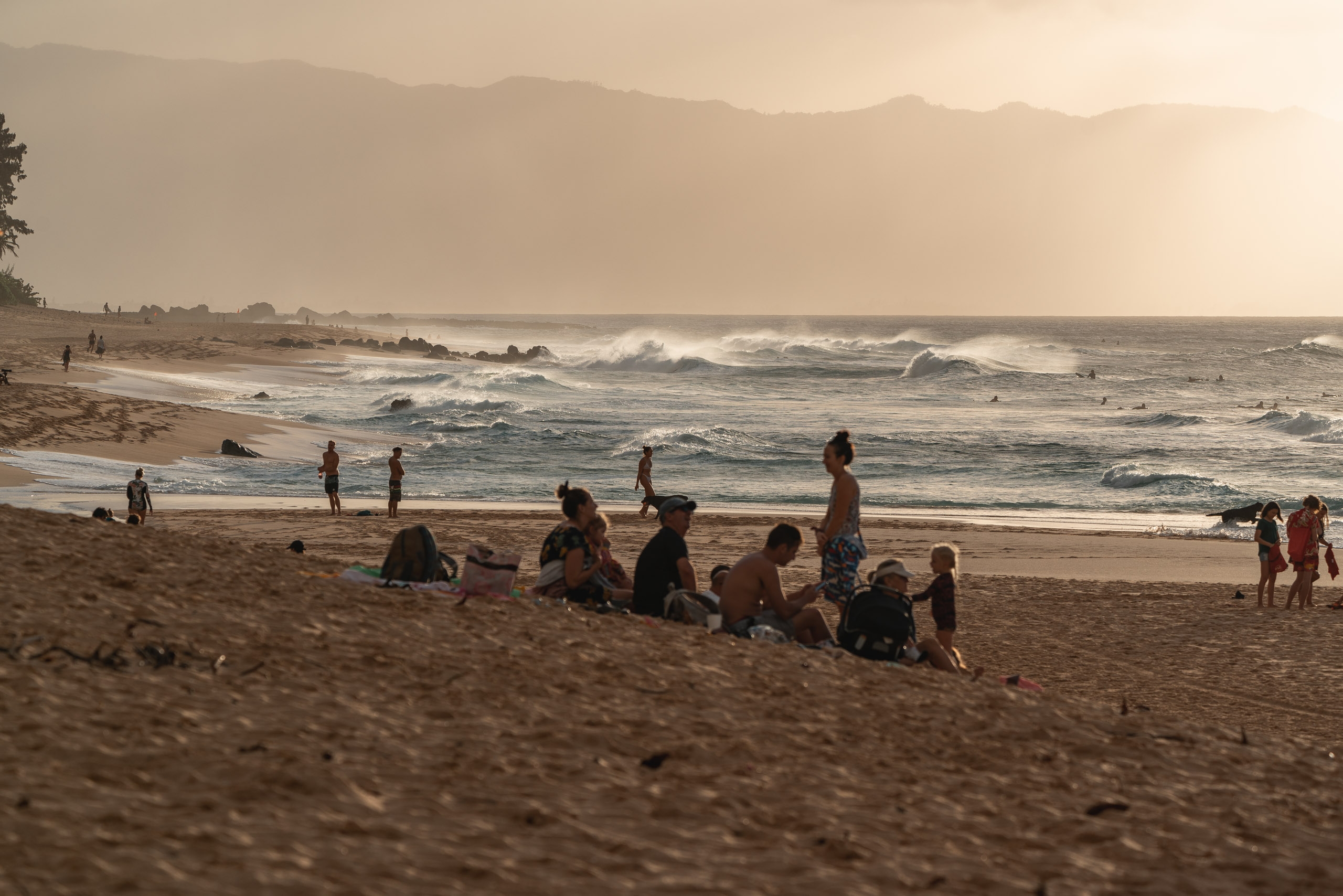 Beachgoers and surfers gather on sandy shore as waves break offshore beneath hazy mountains under golden afternoon light