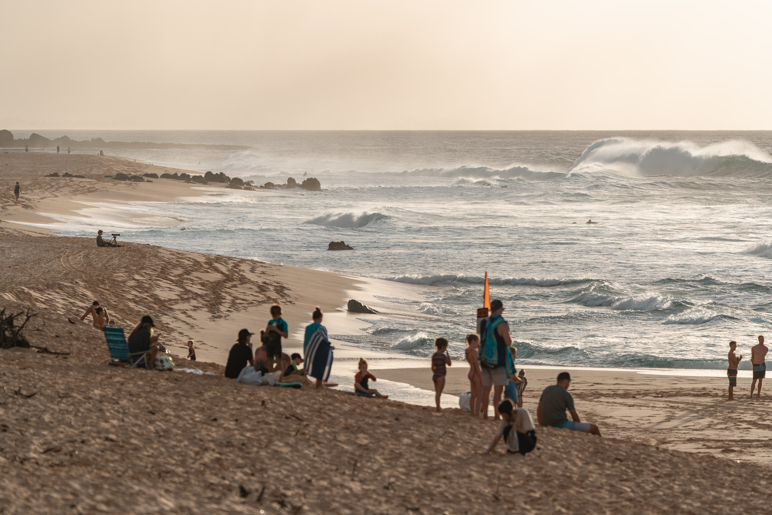 Beachgoers and surfers gather on sandy shores beneath overcast skies as rolling waves crash on Hawaii's North Shore, with distant houses visible along the coast