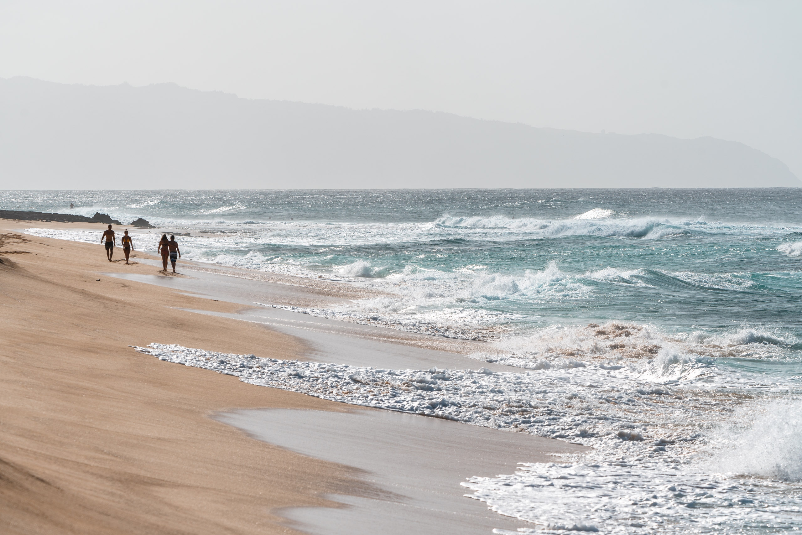 Surfers in wetsuits stand on a sandy beach watching turquoise waves roll in under an overcast sky with distant mountains