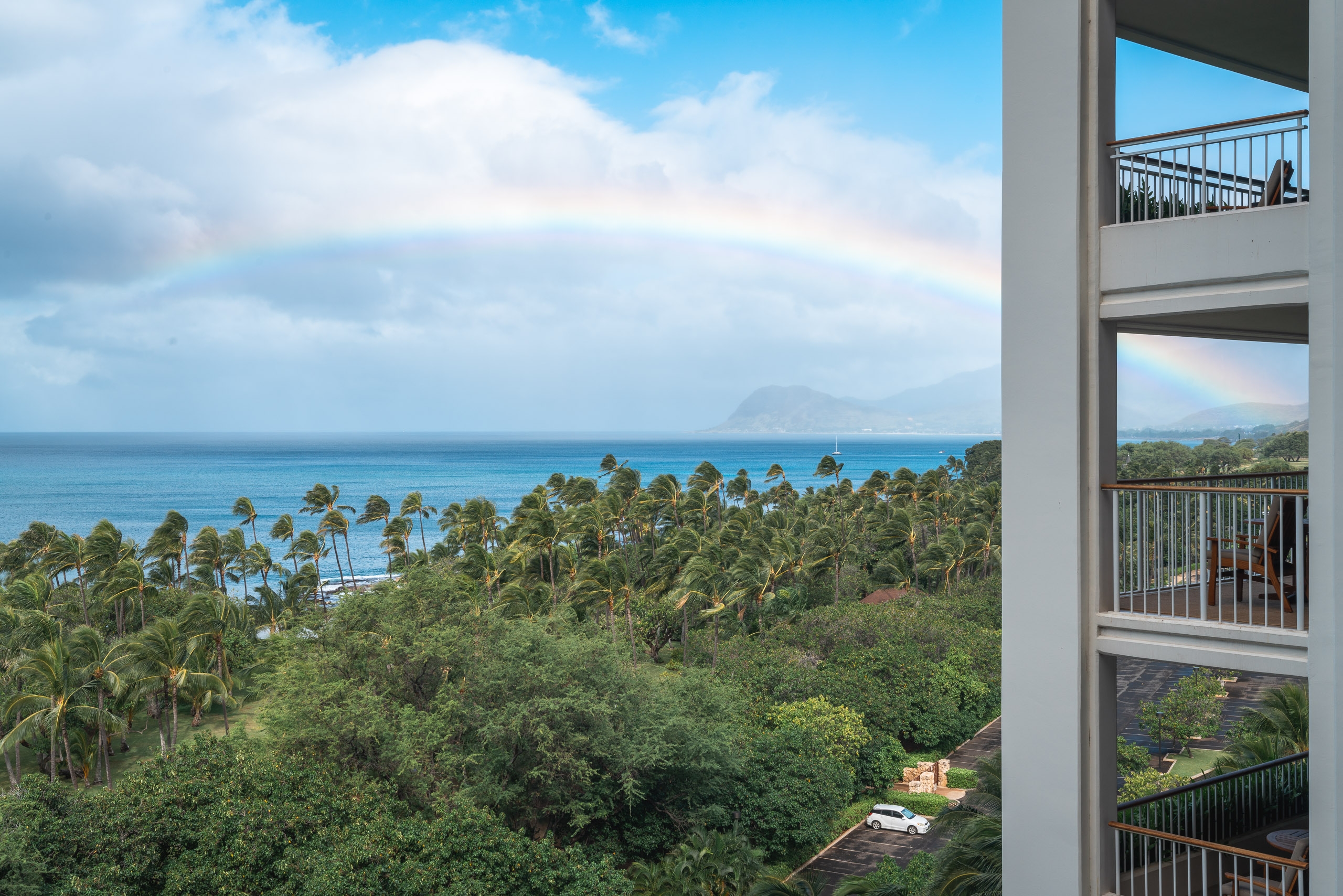 Modern high-rise balcony overlooks a lush tropical landscape with palm trees, turquoise ocean waters, and distant volcanic mountains under partly cloudy skies