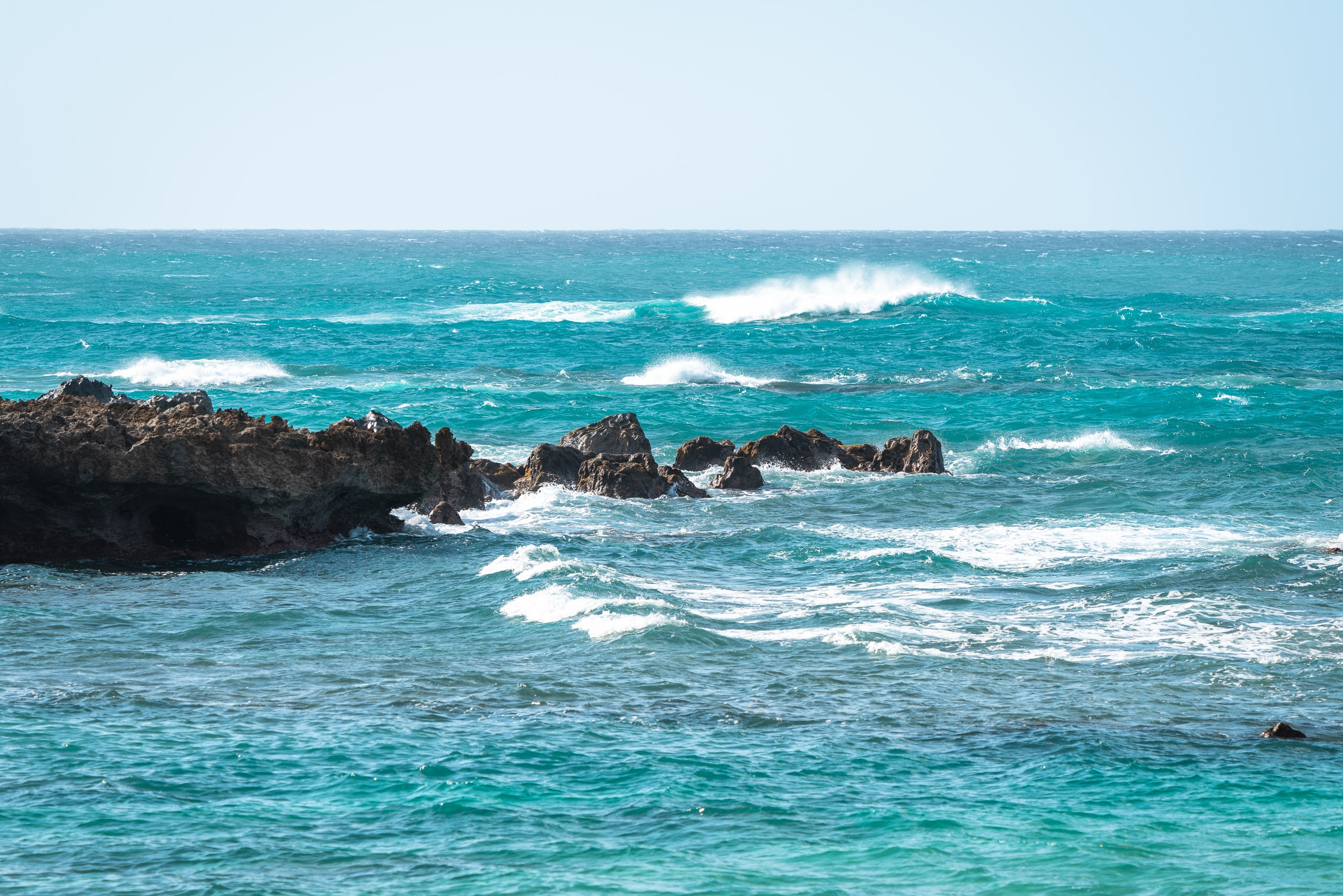 Turquoise waves with white foam crash against dark volcanic rock outcroppings under a clear blue sky on Oahu's North Shore